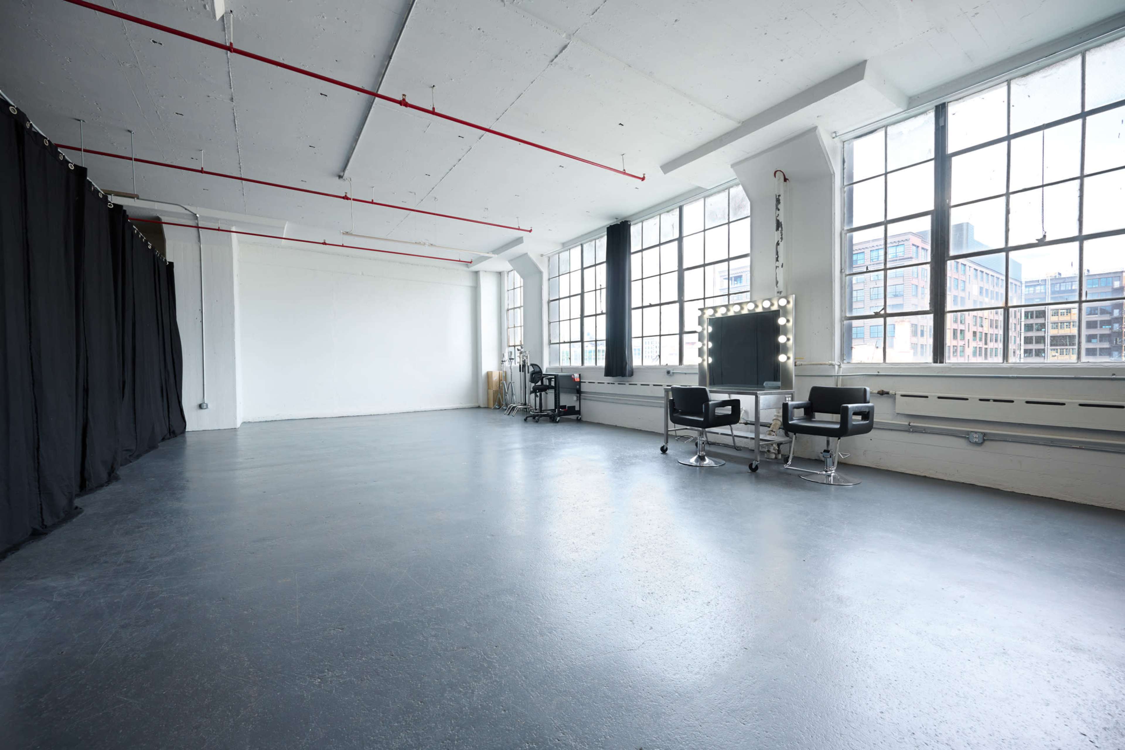 A spacious studio with large windows, black curtains on one side, and two styling chairs next to a mirror with lights.