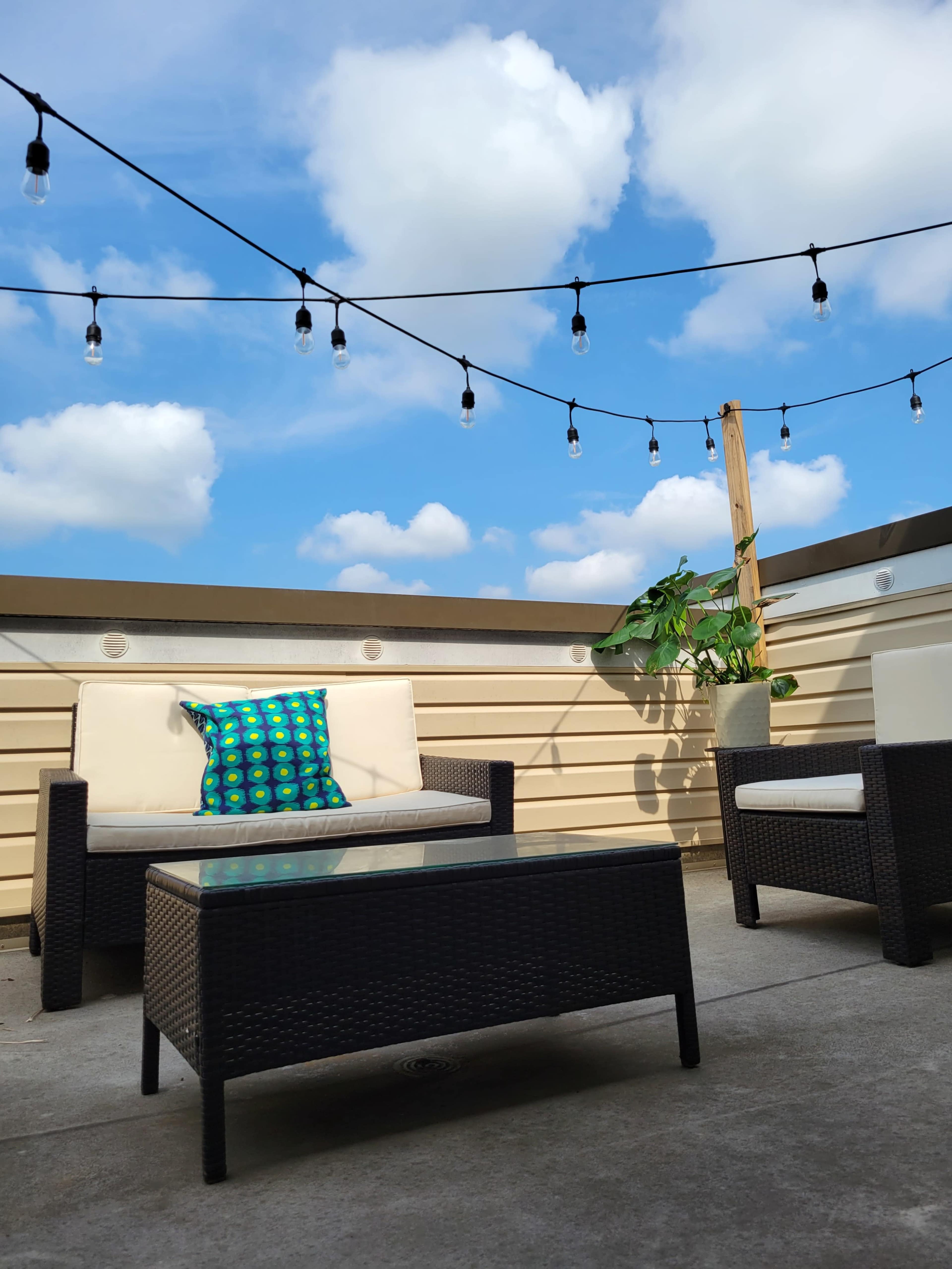 A rooftop patio features two chairs and a table, with a colorful pillow on one chair and string lights overhead against a backdrop of blue sky and clouds.