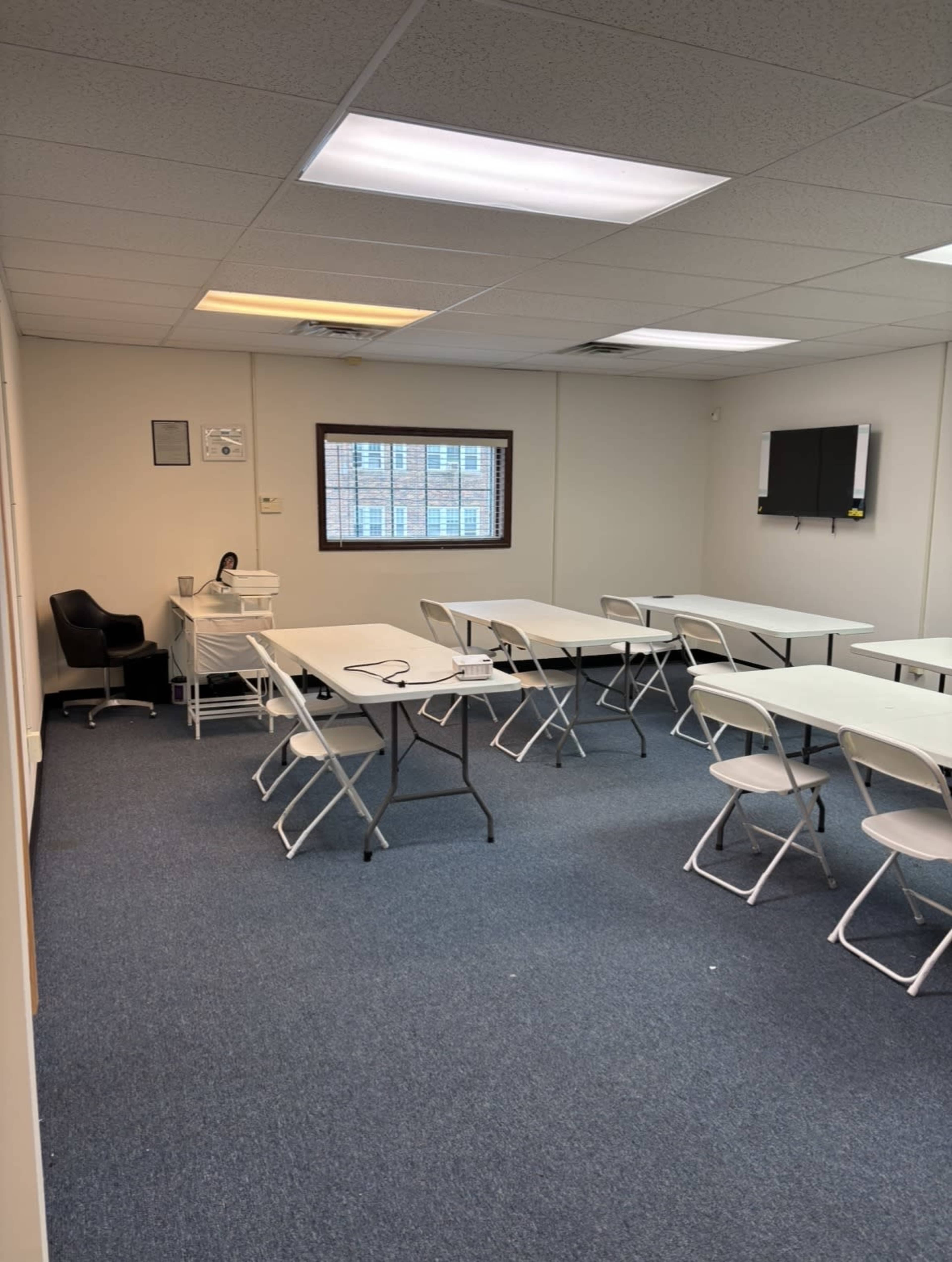 The image shows a sparsely furnished classroom with multiple folding tables and chairs arranged in rows, a desk in the corner, and a window letting in natural light.