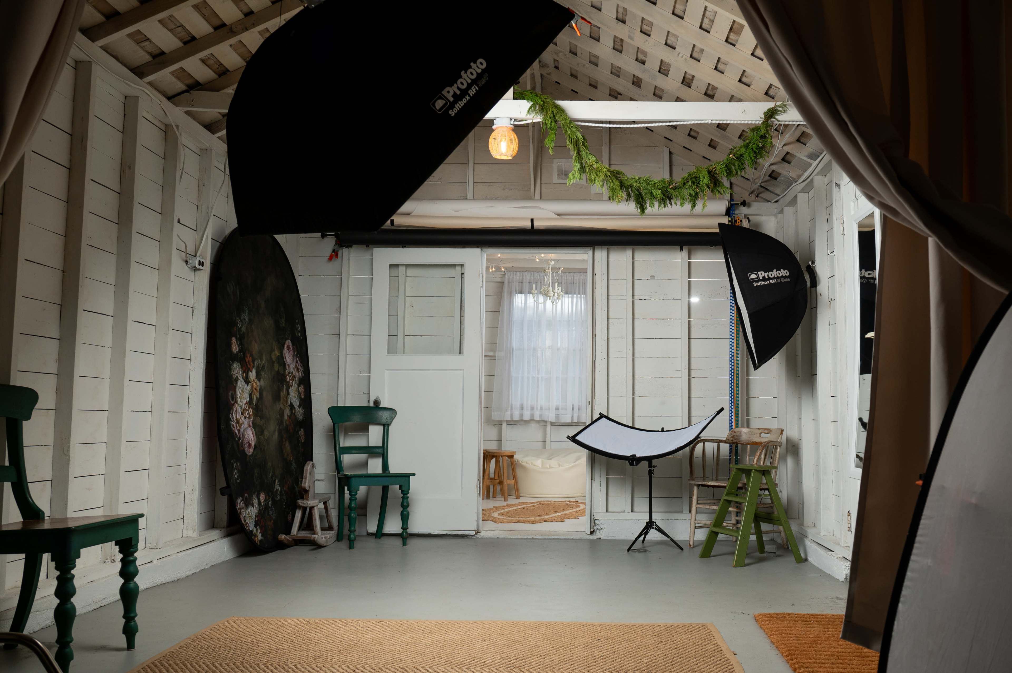 The image shows a photography studio with lighting equipment, green chairs, and a backdrop set up in a white wooden room.