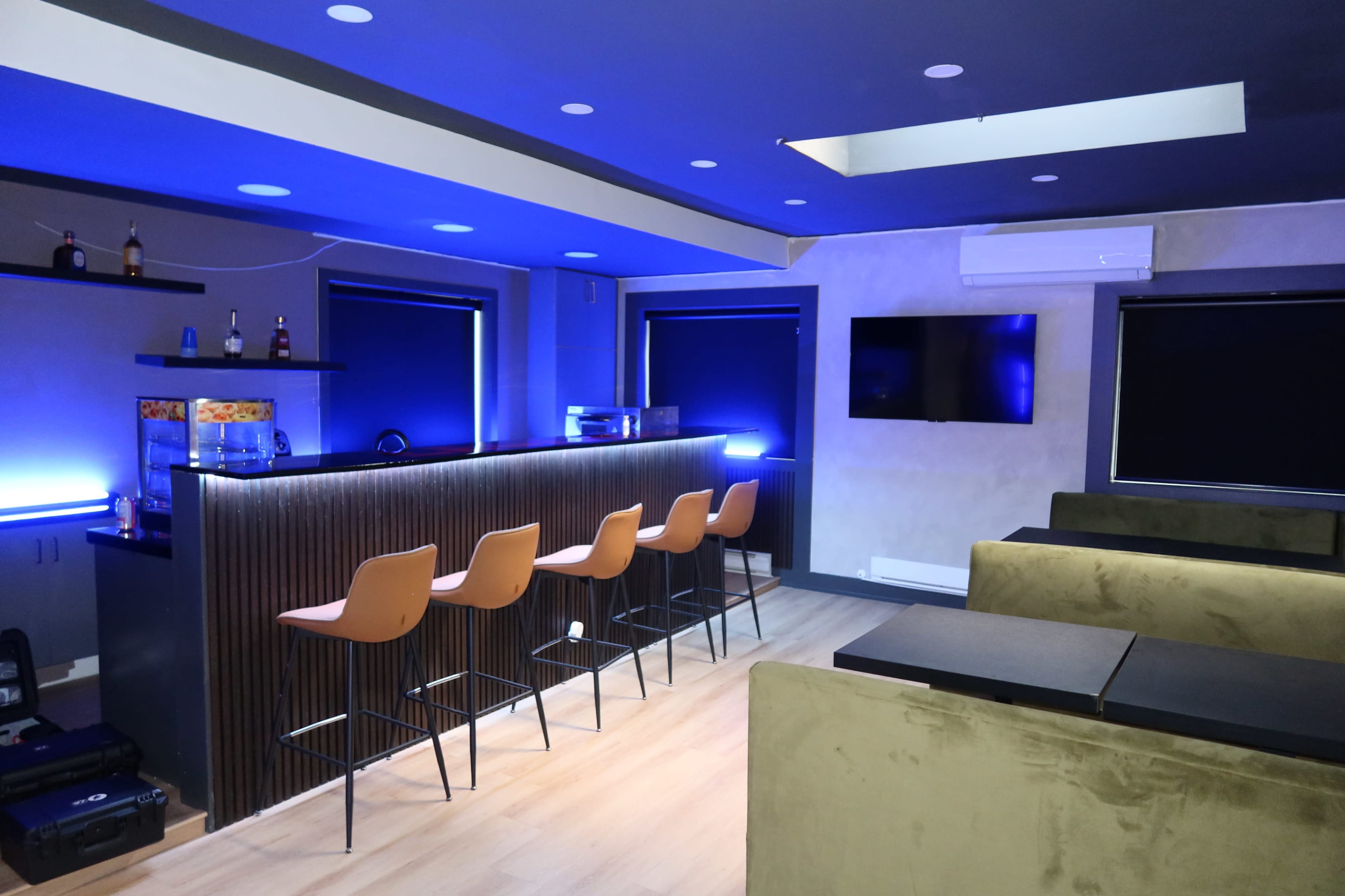The image shows a modern bar area with a sleek counter, several high stools, and a wall-mounted television, illuminated by blue lighting.