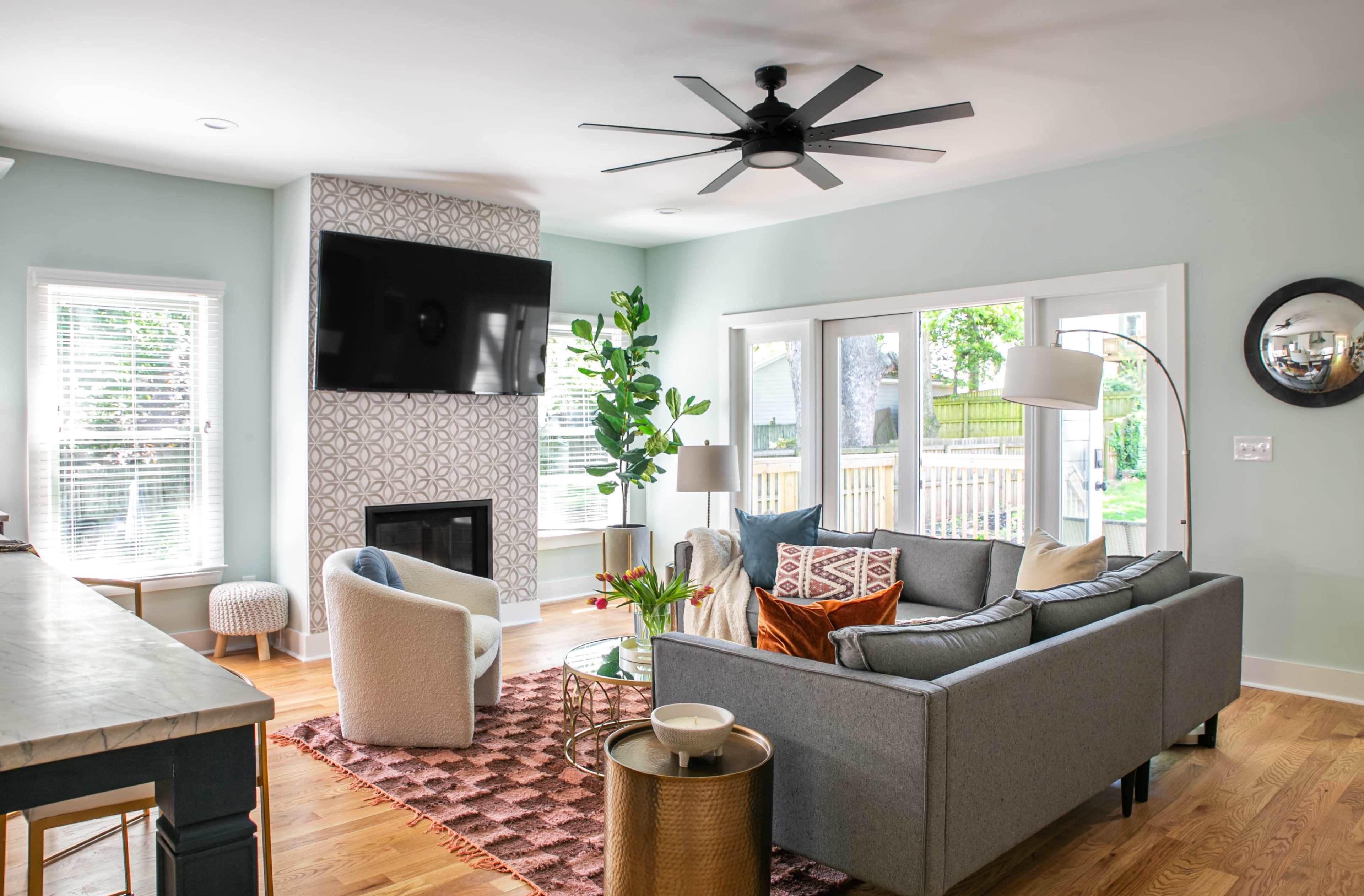 A modern living room features a gray sectional sofa, a large television mounted on a patterned wall, and large windows that provide natural light.