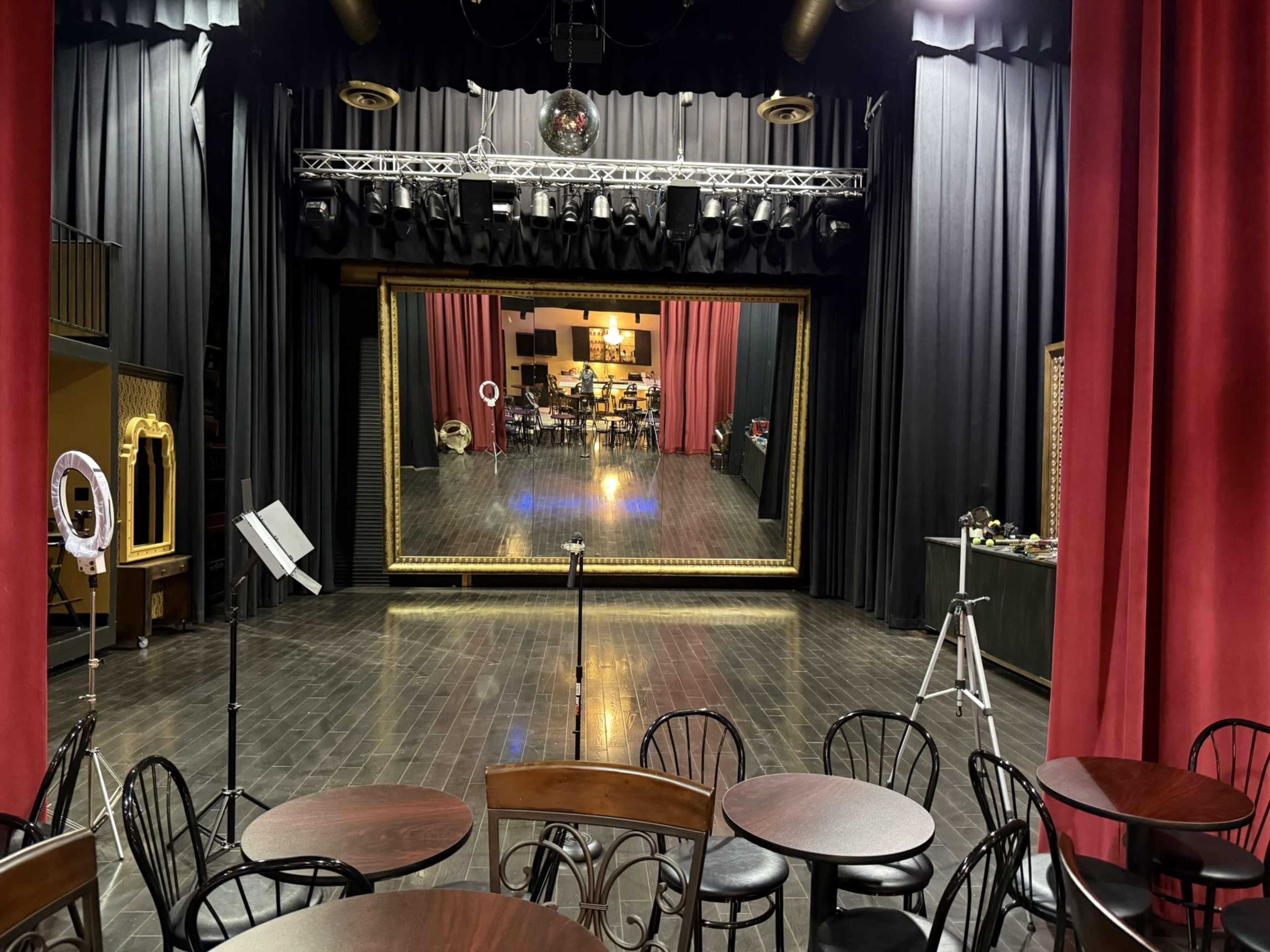 The image shows a stage with a large mirror framed in gold, surrounded by red curtains, and several tables and chairs set up in the foreground.