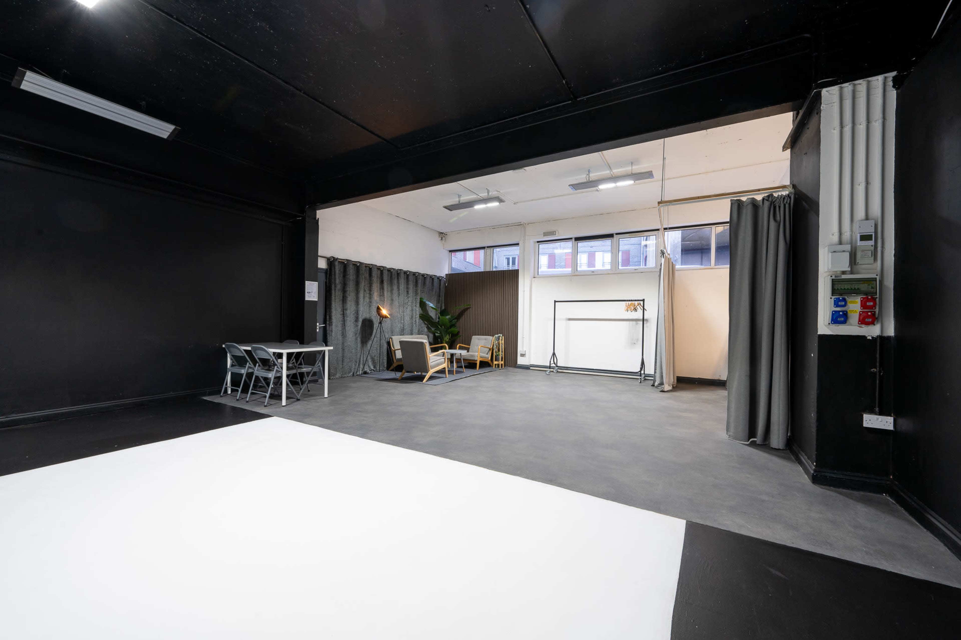 The image shows a spacious studio with black walls, a gray floor, a white section on the ground, a table with chairs, and a seating area featuring two chairs and a plant.