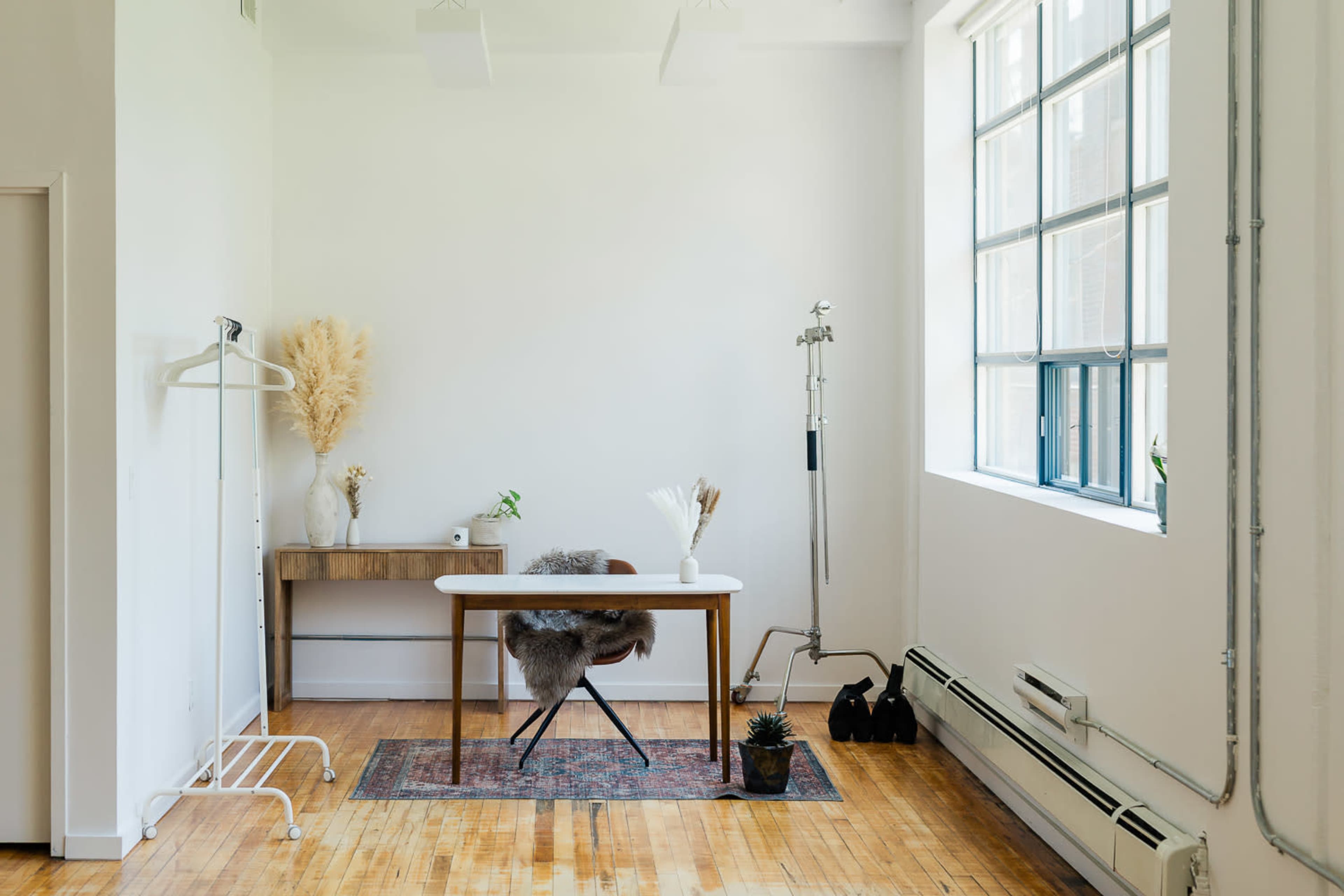 The image shows a minimalist workspace featuring a wooden desk, a hanging clothing rack, and large windows allowing natural light to fill the room.