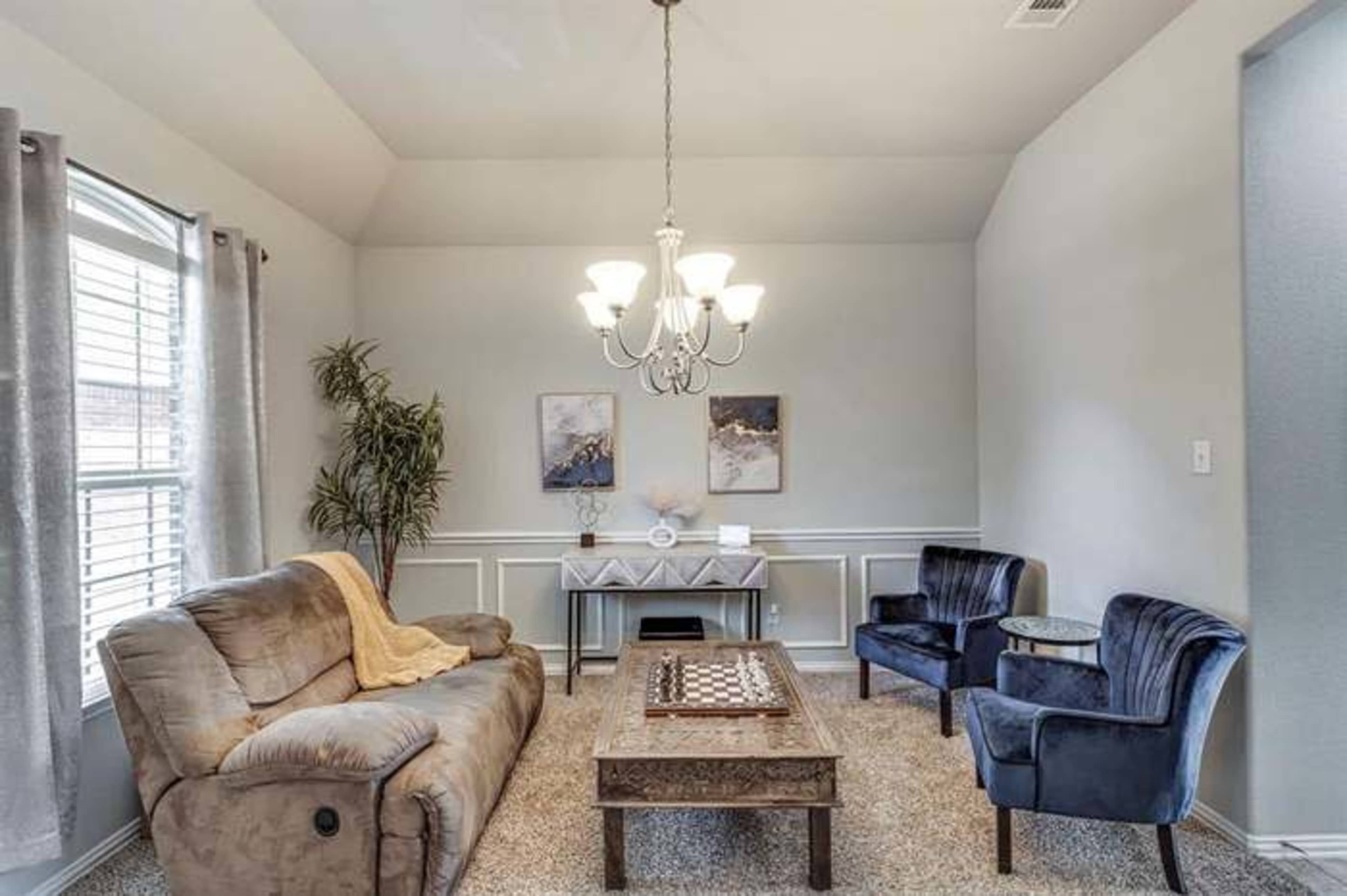 The image shows a modern living room with a beige sofa, two blue upholstered chairs, a wooden coffee table, and a chandelier.