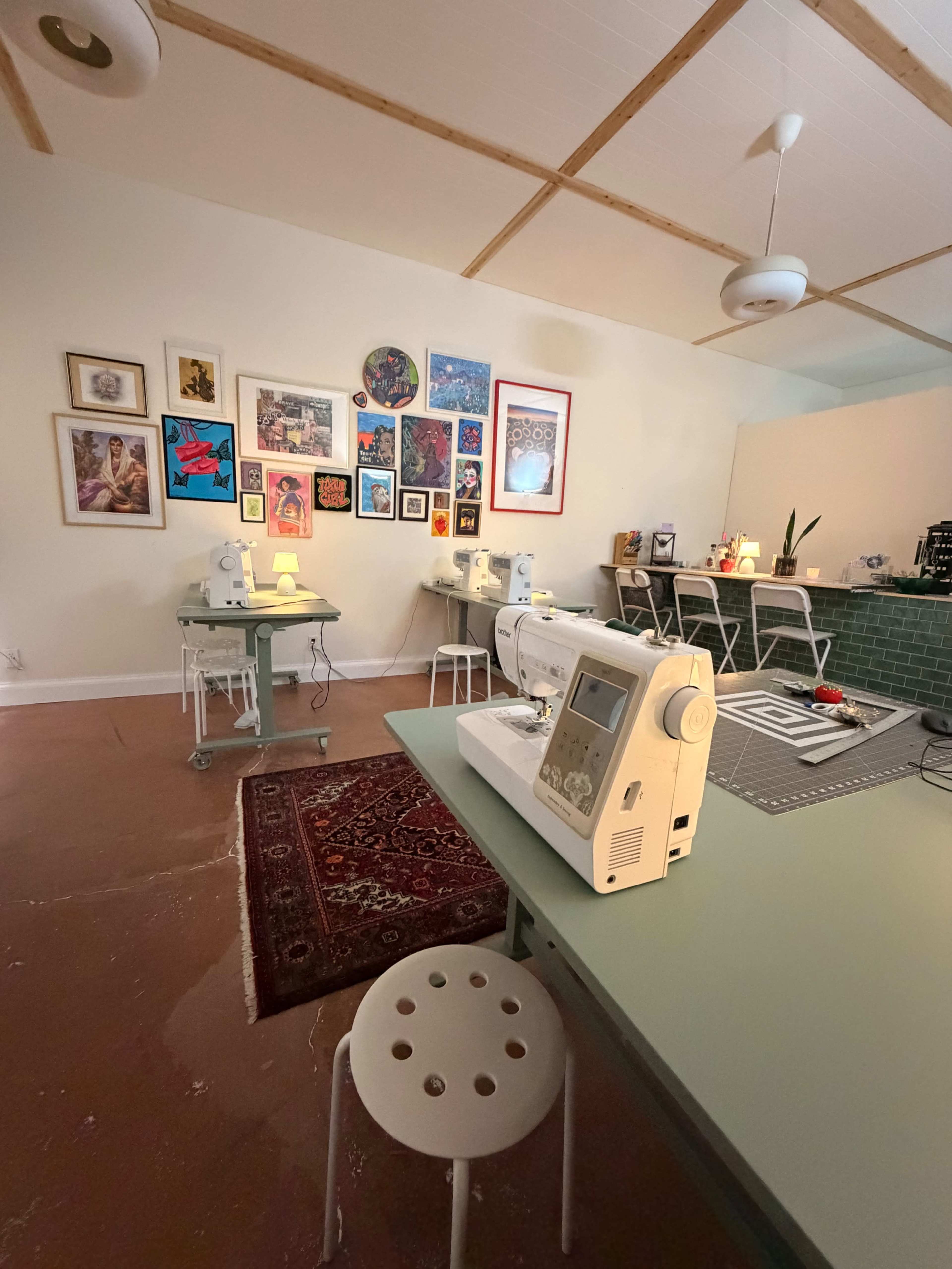 The image shows a bright sewing studio featuring multiple sewing machines on green tables, surrounded by colorful art on the wall and a decorative rug on the floor.