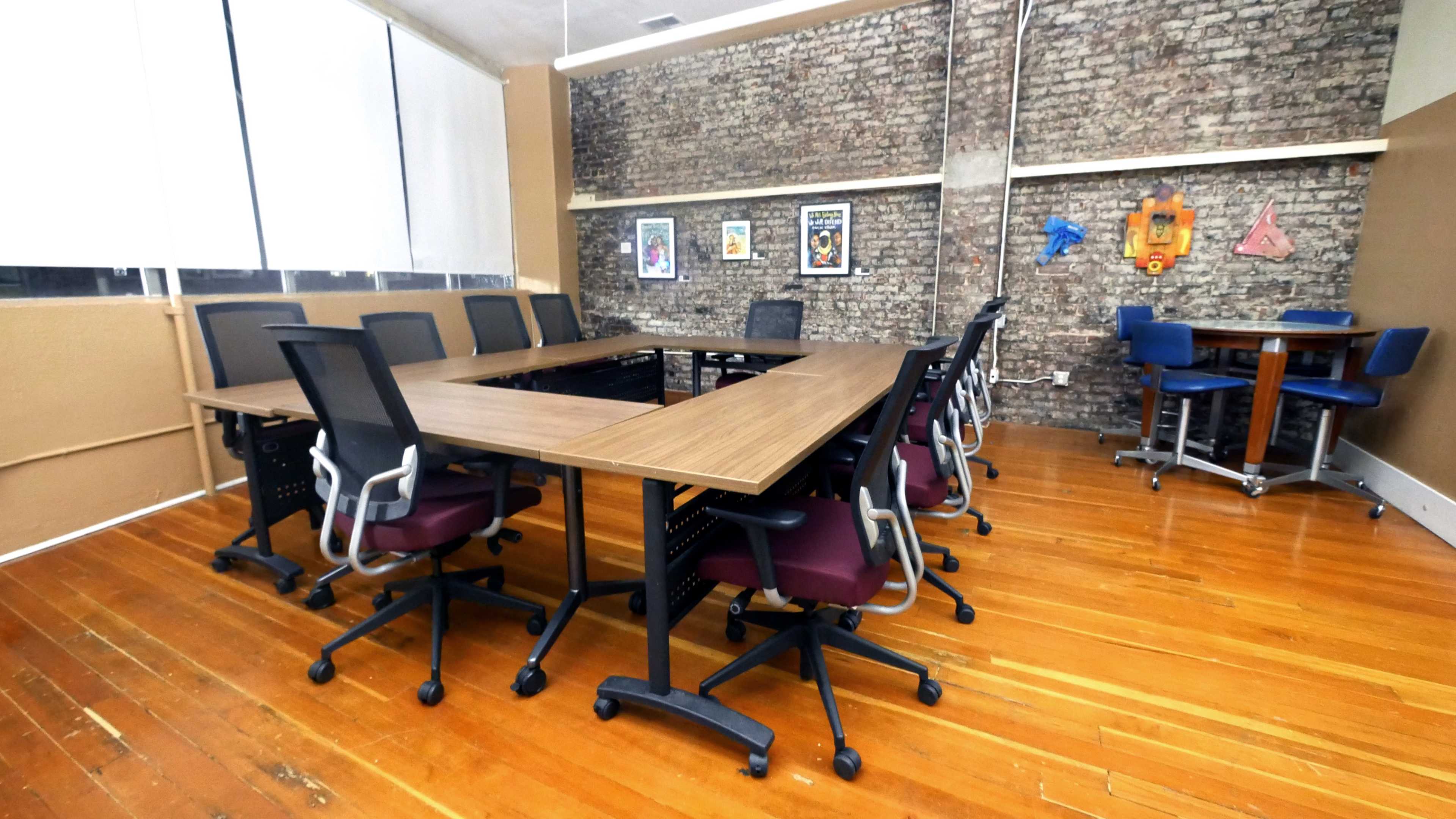 A conference room features a large rectangular table surrounded by office chairs, with brick walls and various artworks displayed.