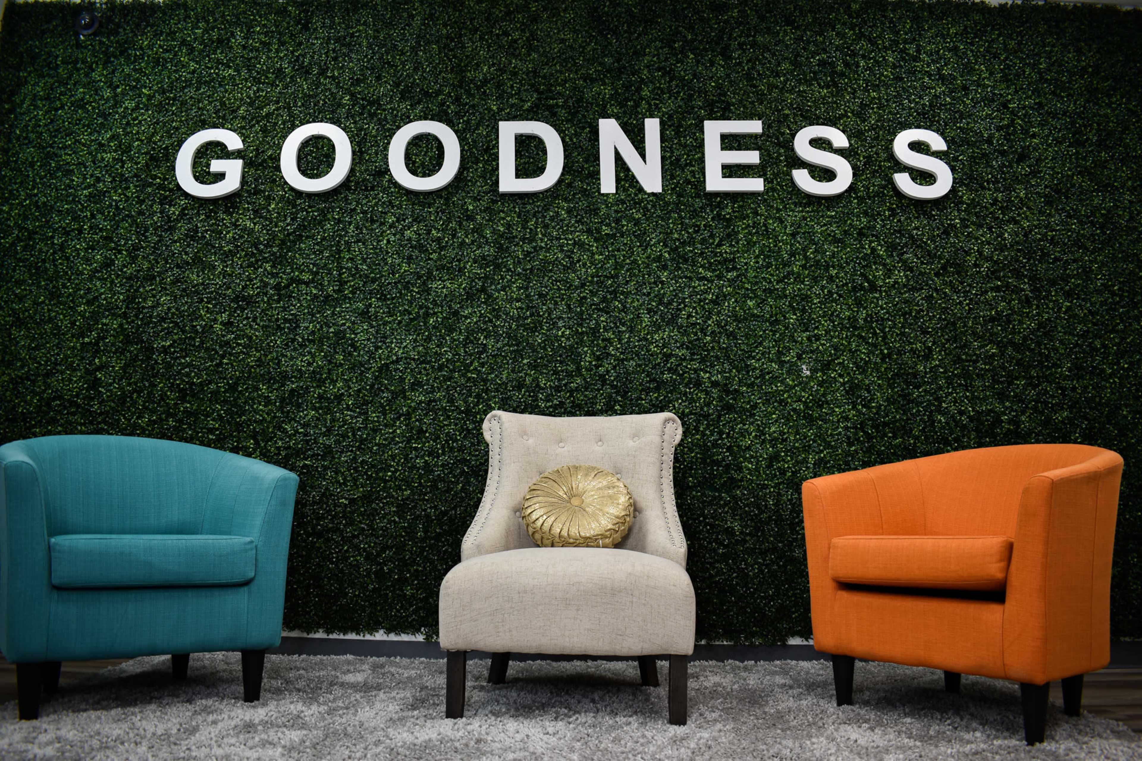 Two armchairs, one turquoise and one orange, facing a beige chair with a decorative cushion, against a green wall with the word "GOODNESS" prominently displayed.