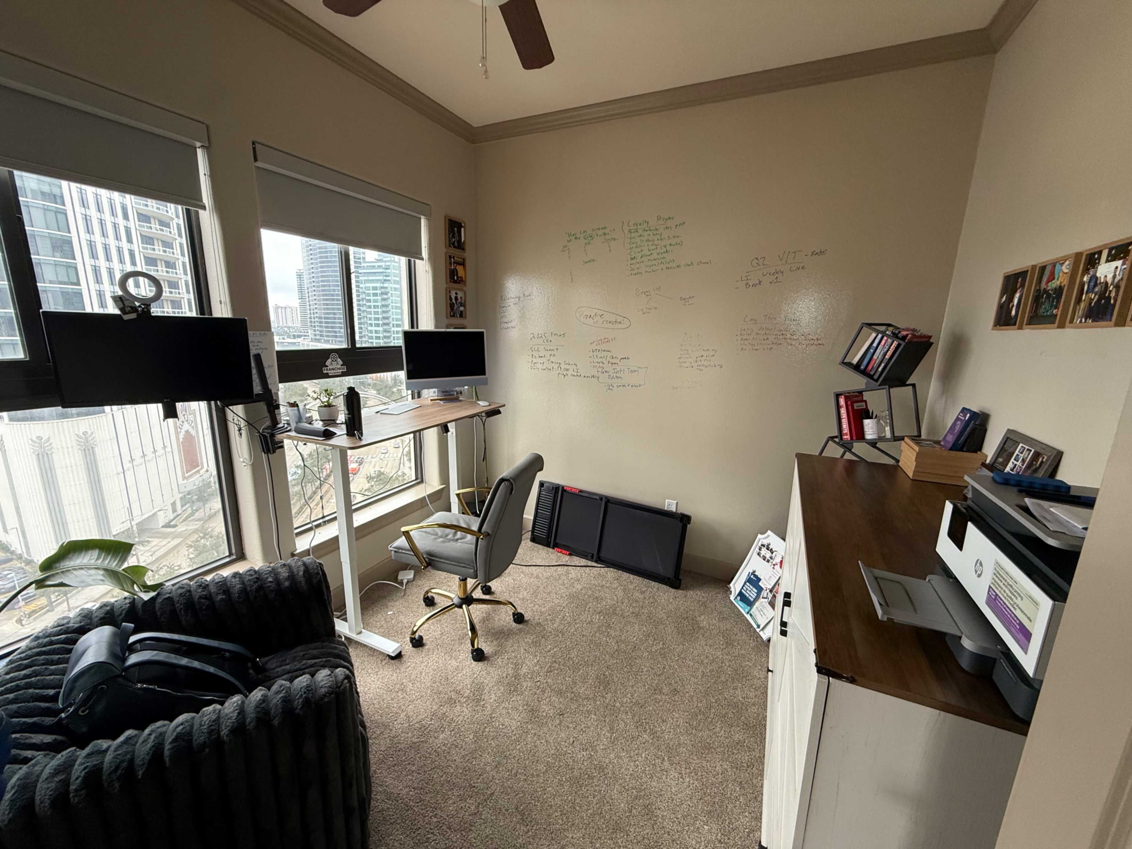 The image shows a home office space featuring a standing desk, two computer monitors, a comfortable chair, a wall covered with handwritten notes, and a storage unit with books and documents.