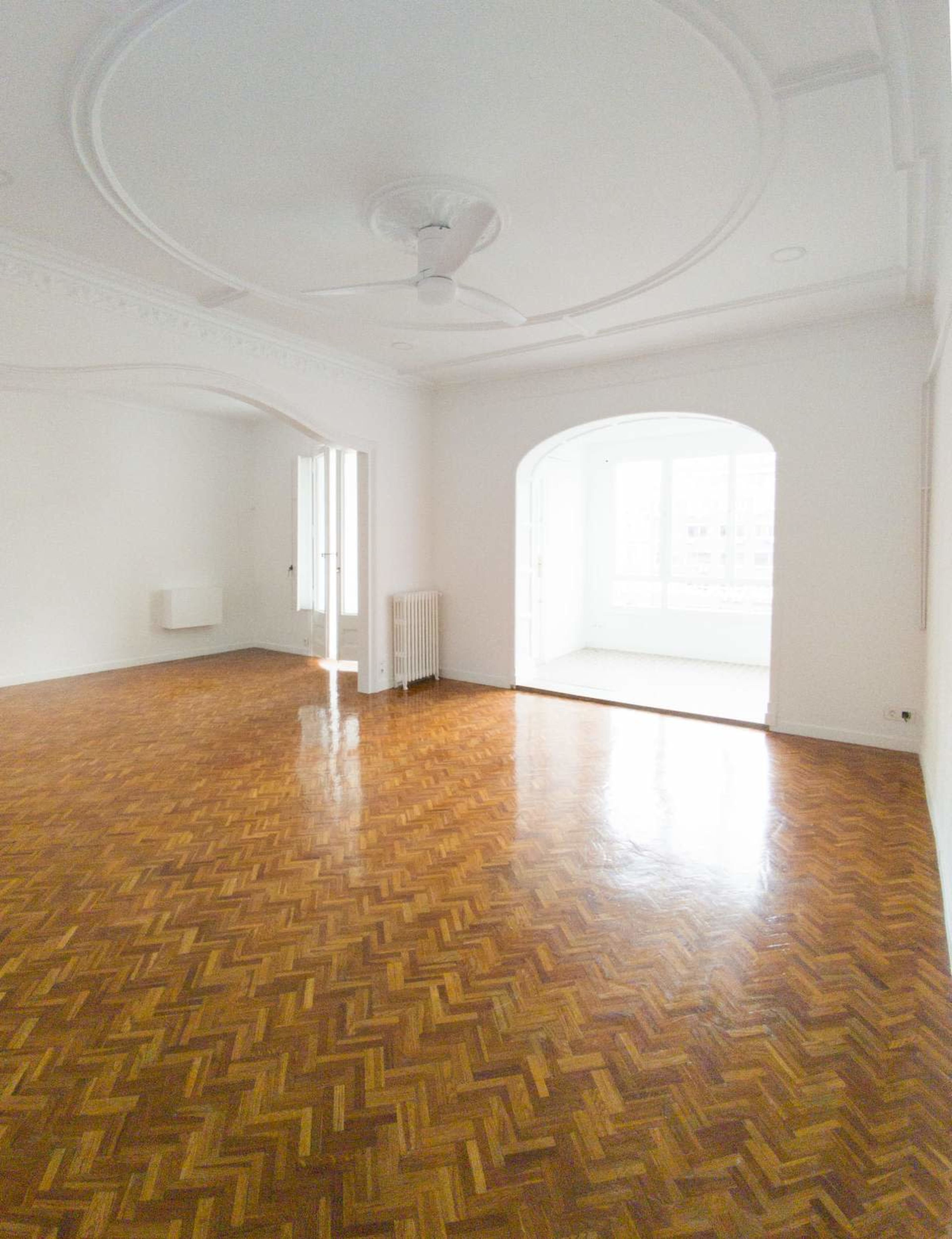 The image shows a spacious, airy room with a herringbone-patterned wooden floor and large arched doorways leading to another well-lit area.