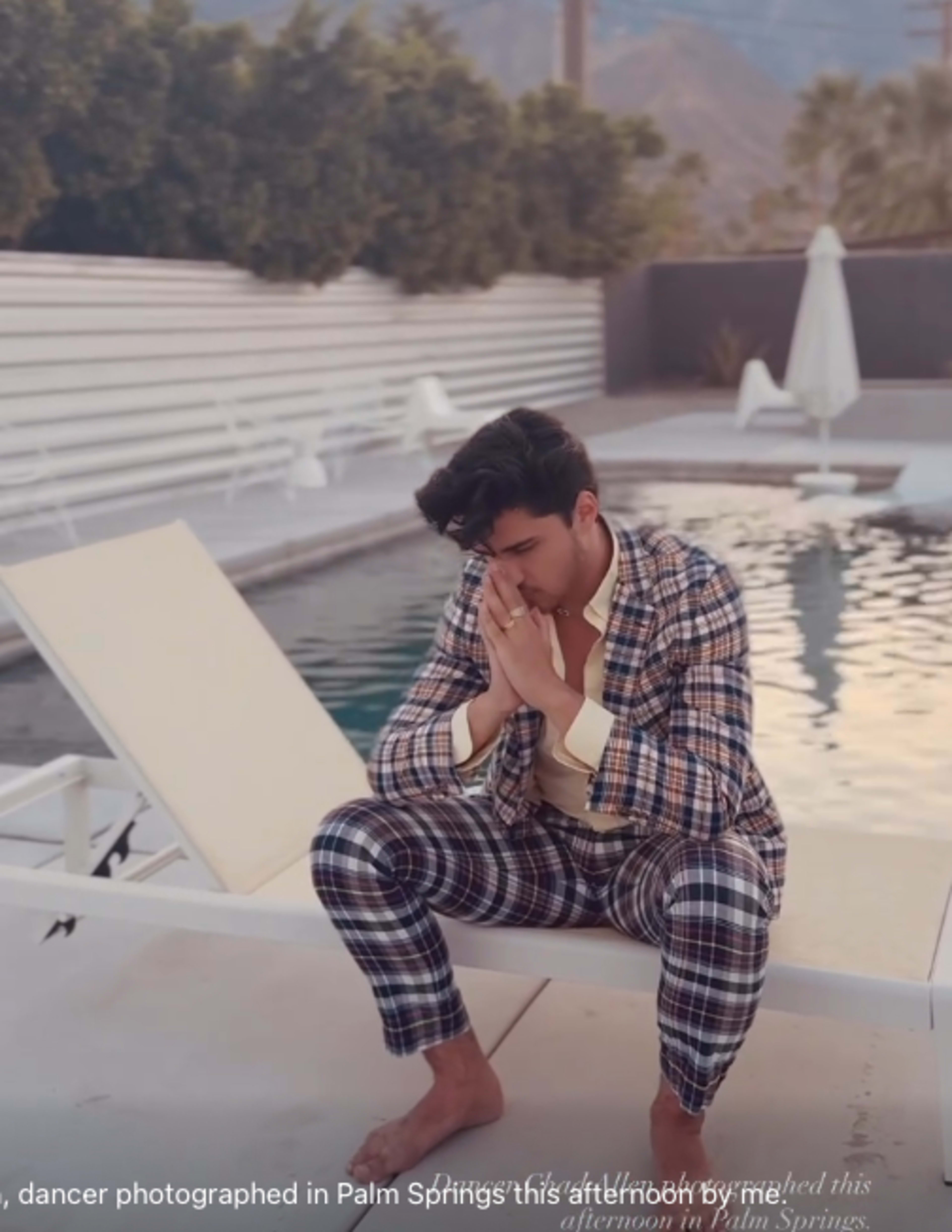 A man in a patterned suit sits by a pool, resting his elbows on his knees with his hands clasped in front of him.
