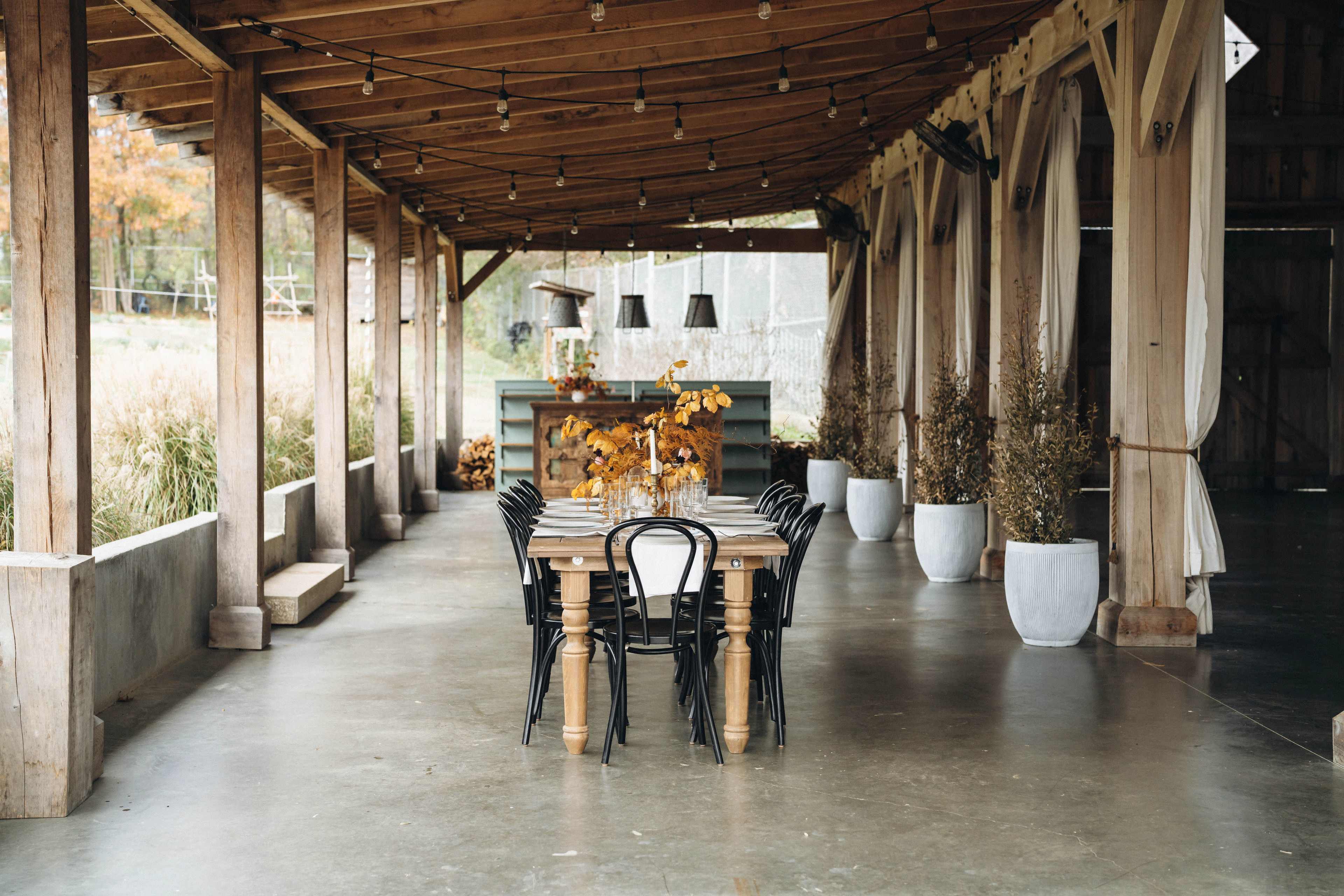 A long wooden table is set with black chairs beneath a covered porch adorned with hanging lights and potted plants.