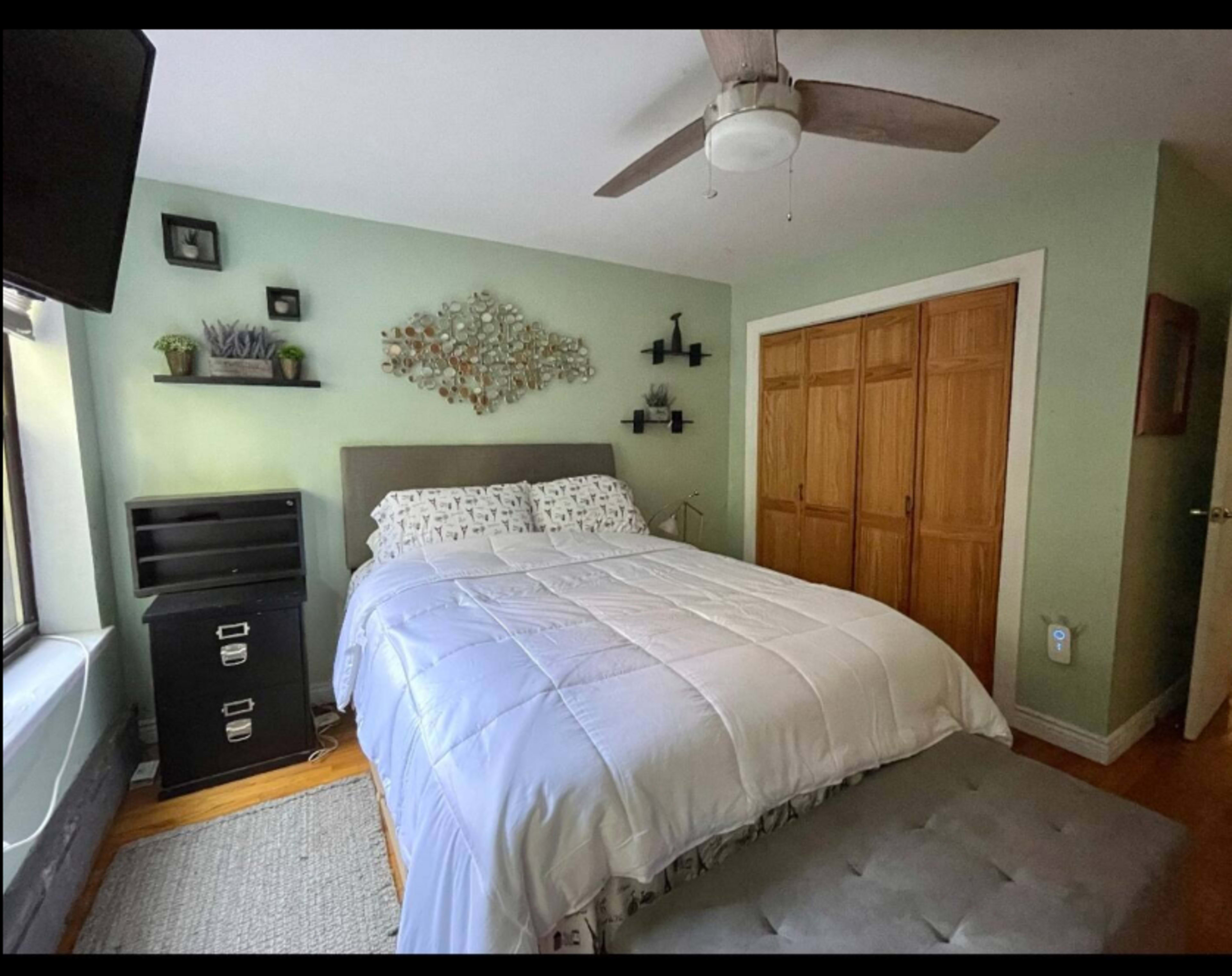 A small bedroom featuring a bed with white bedding, a wall-mounted decorative piece, a ceiling fan, and wooden closets.