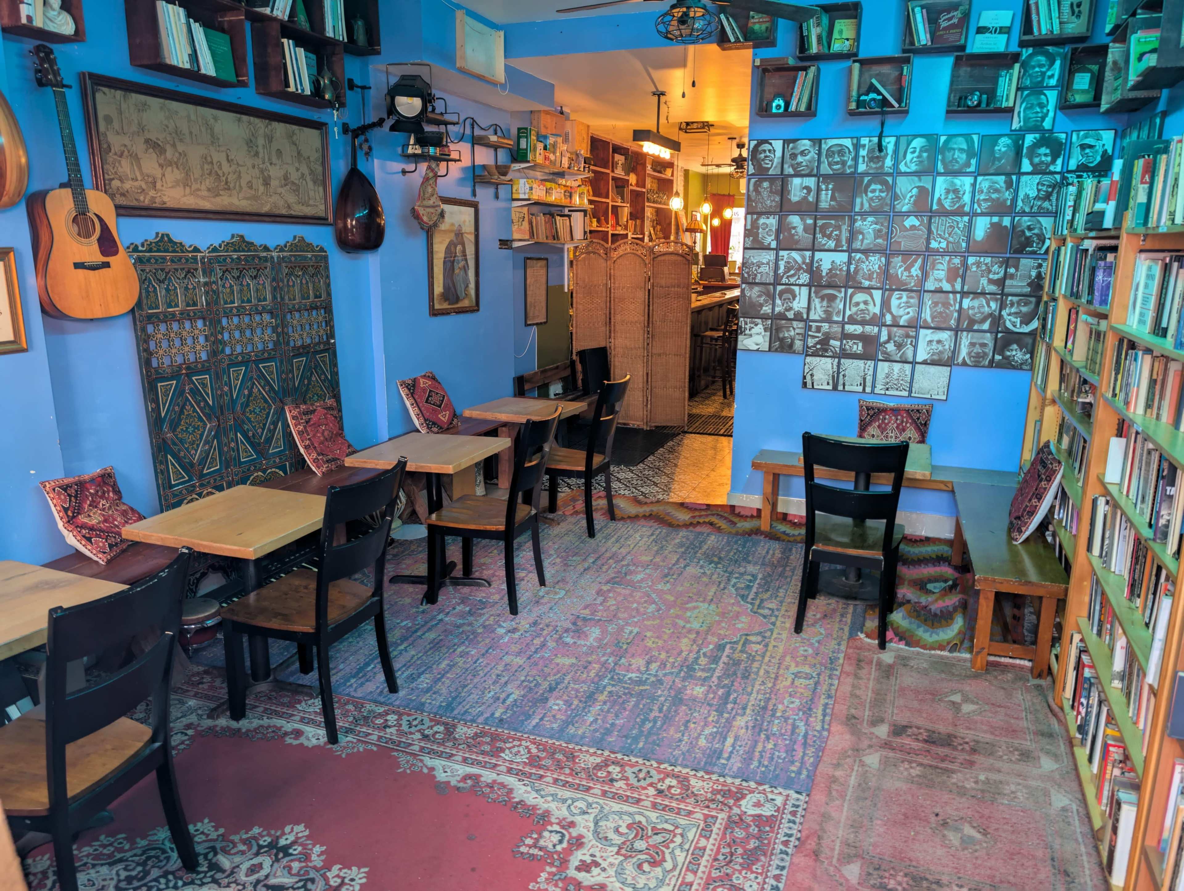 The image shows a cozy café with wooden tables and chairs arranged on colorful rugs, lined with bookshelves and adorned with cultural decor.