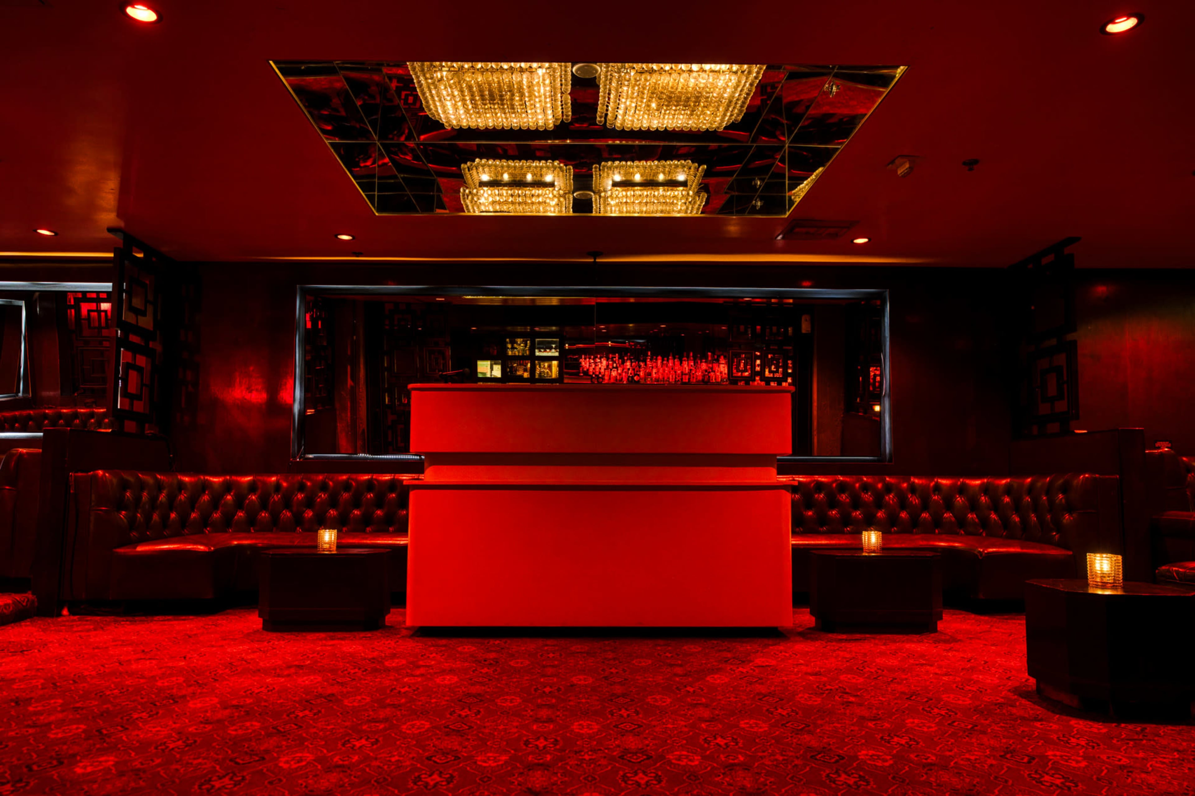 A dimly lit lounge features red walls, tufted seating, and a mirrored ceiling, creating an intimate atmosphere.