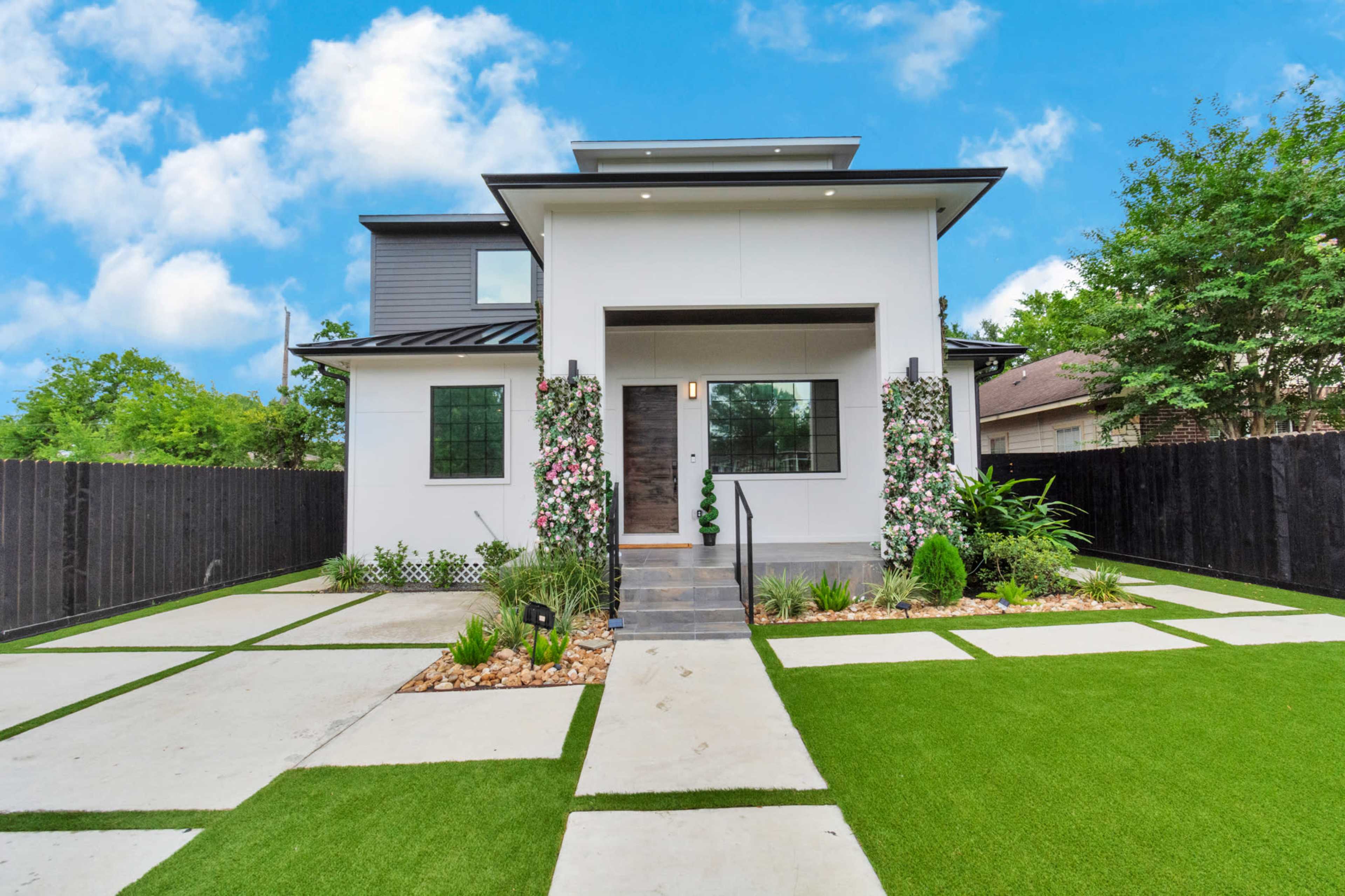 A modern two-story house with a gray and white facade features a front walkway lined with greenery and flowering plants.