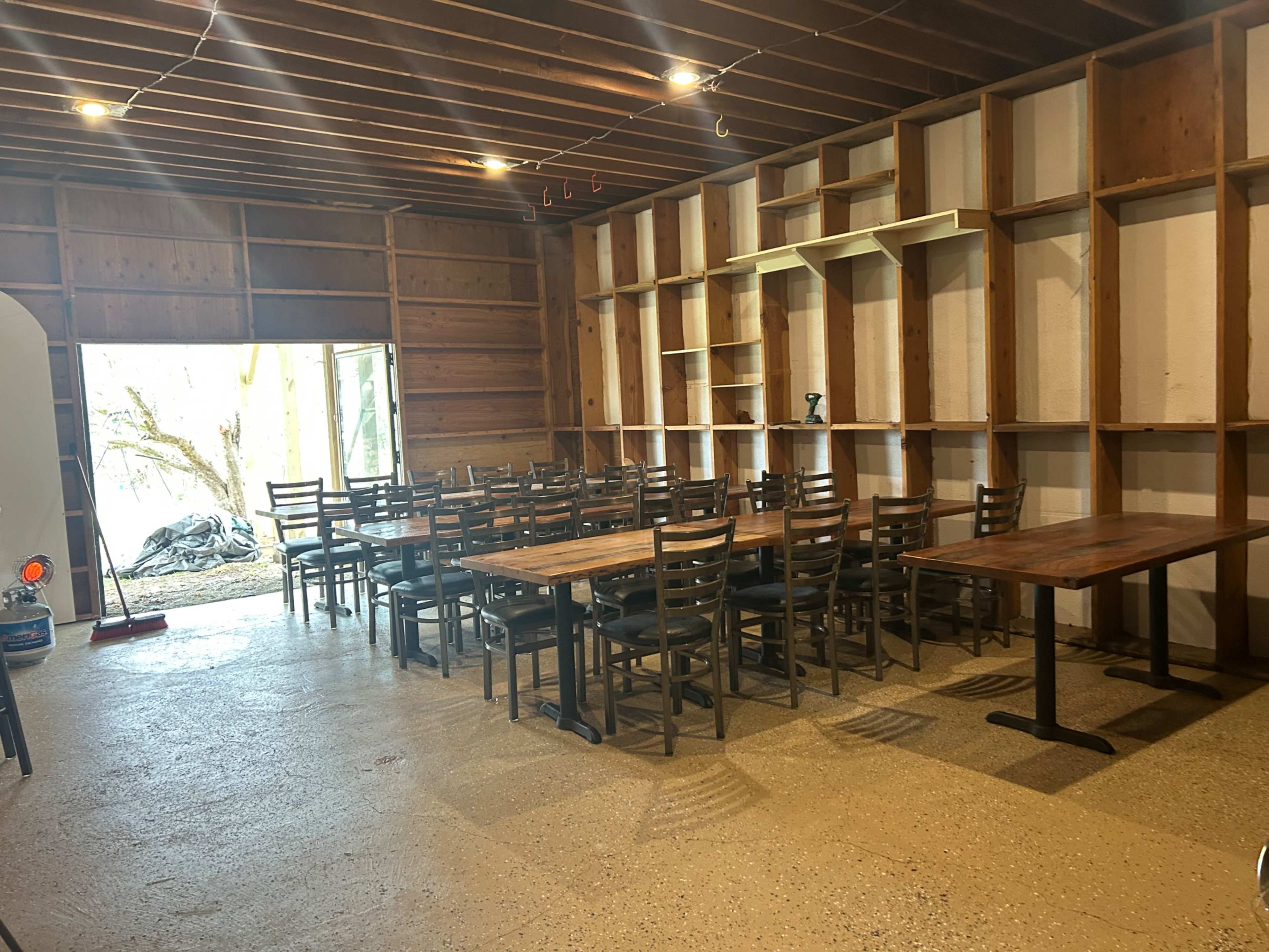 The image shows a spacious, unfinished interior room with multiple wooden tables and chairs arranged for dining, and an open entry leading outside.