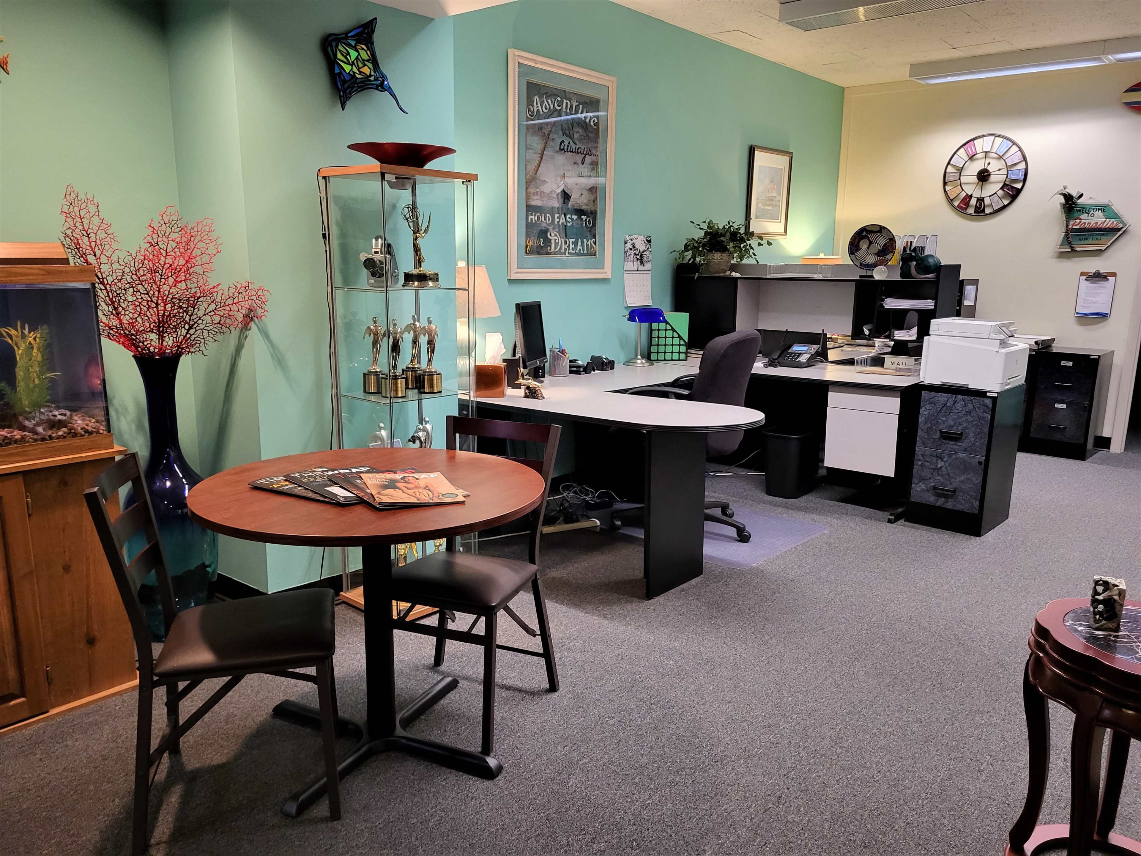 The image shows an office space featuring a circular table with two chairs, a fish tank, a display cabinet with trophies, and a desk area with office equipment.