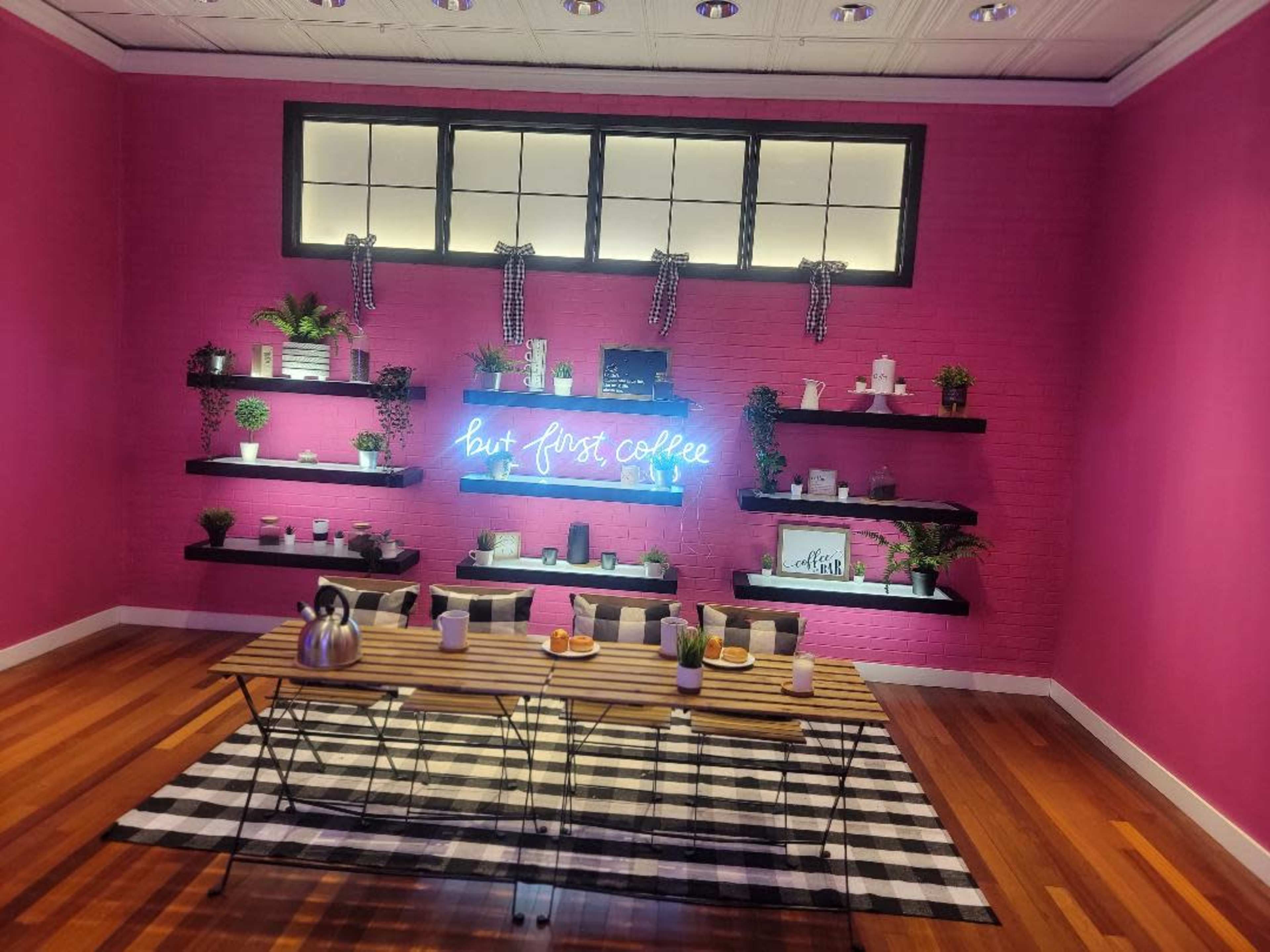 A vibrant pink room with shelves displaying plants and decorative items, and a neon sign that reads "but first coffee" above a table set with coffee mugs and a table runner.