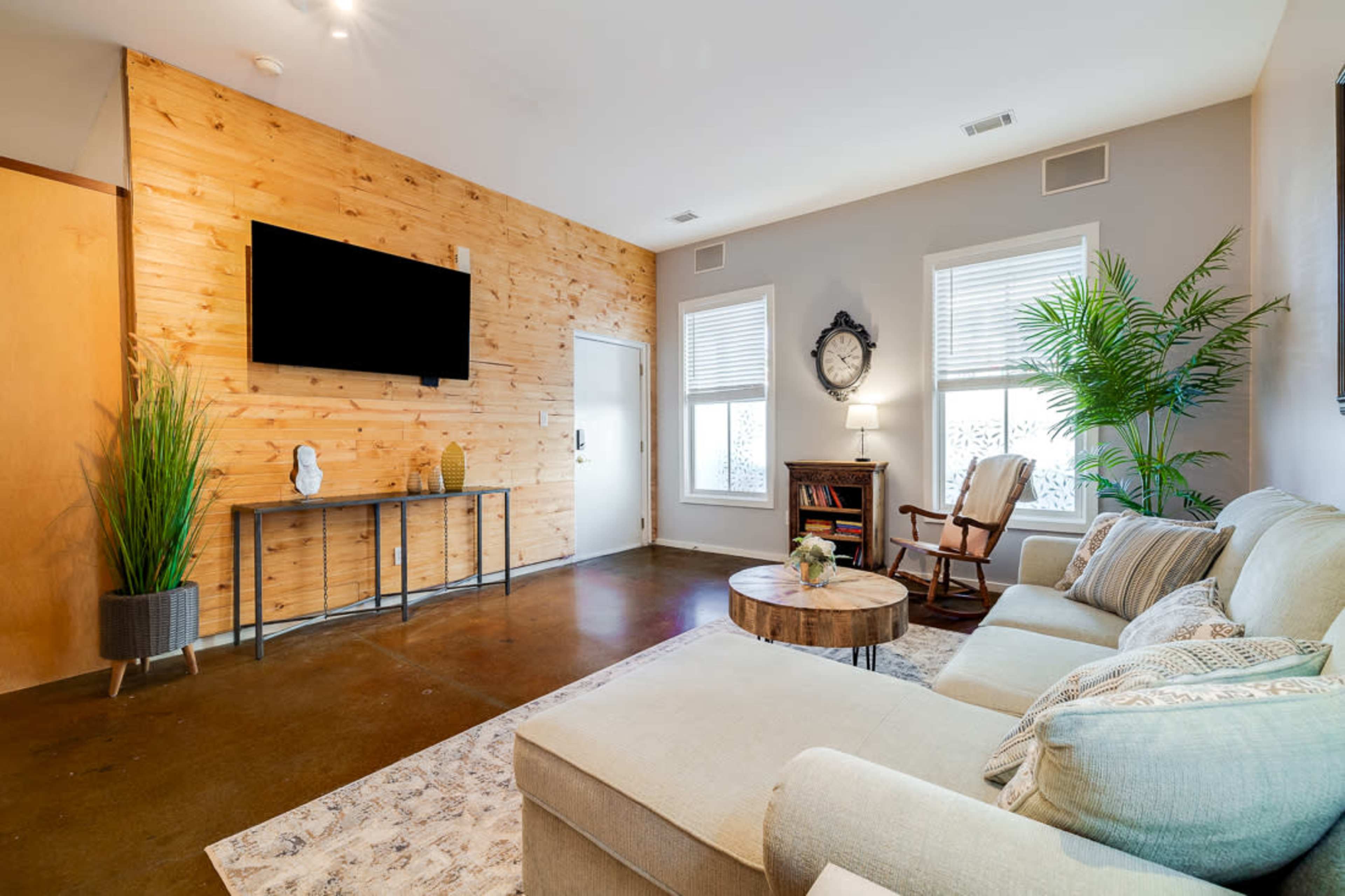 The image shows a living room featuring a beige sectional sofa, a wooden accent wall, a round table, and a large television mounted on the wall.
