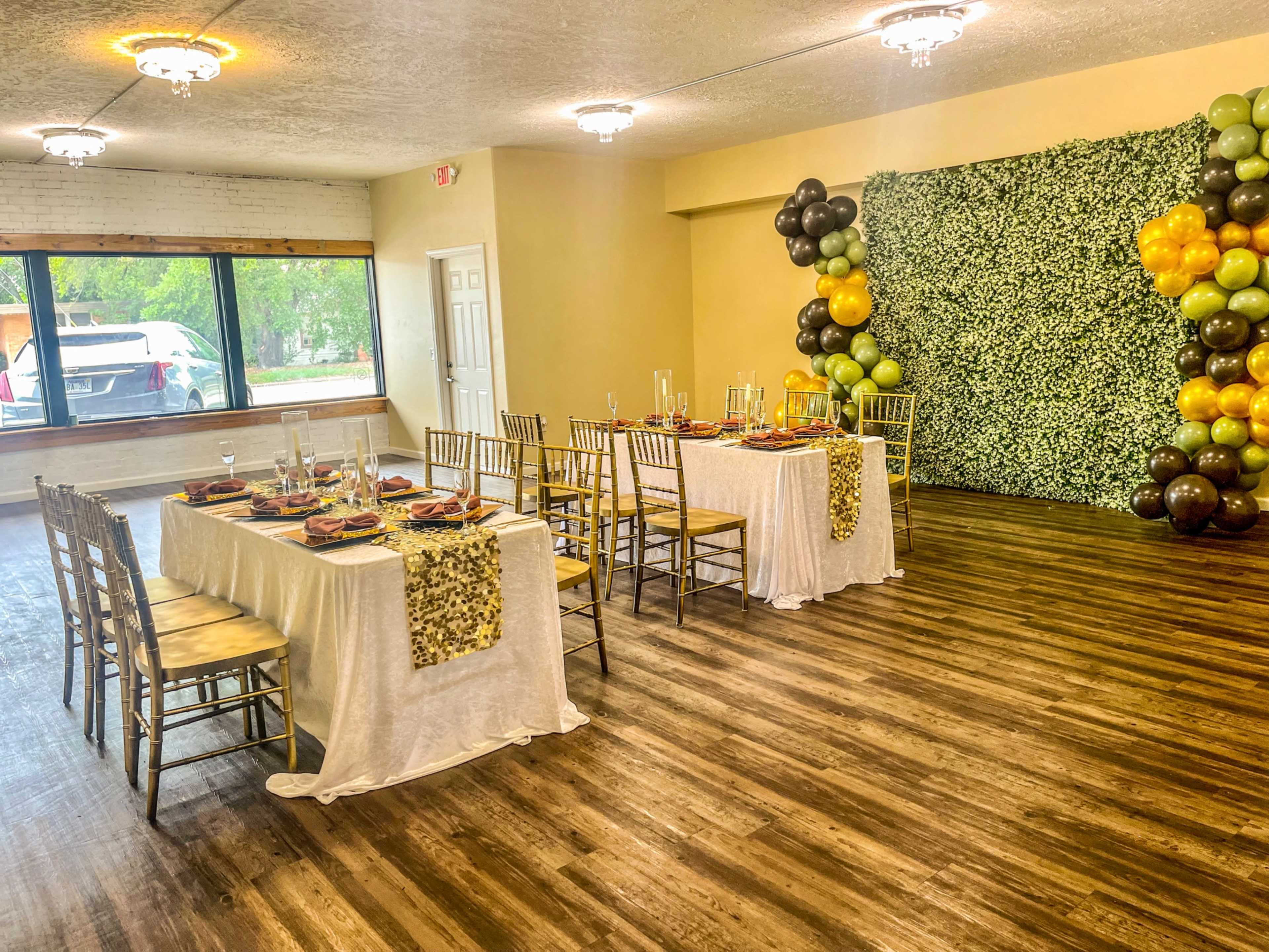 A decorated dining area features two tables set with white tablecloths, elegant place settings, and a backdrop of green and gold balloons.