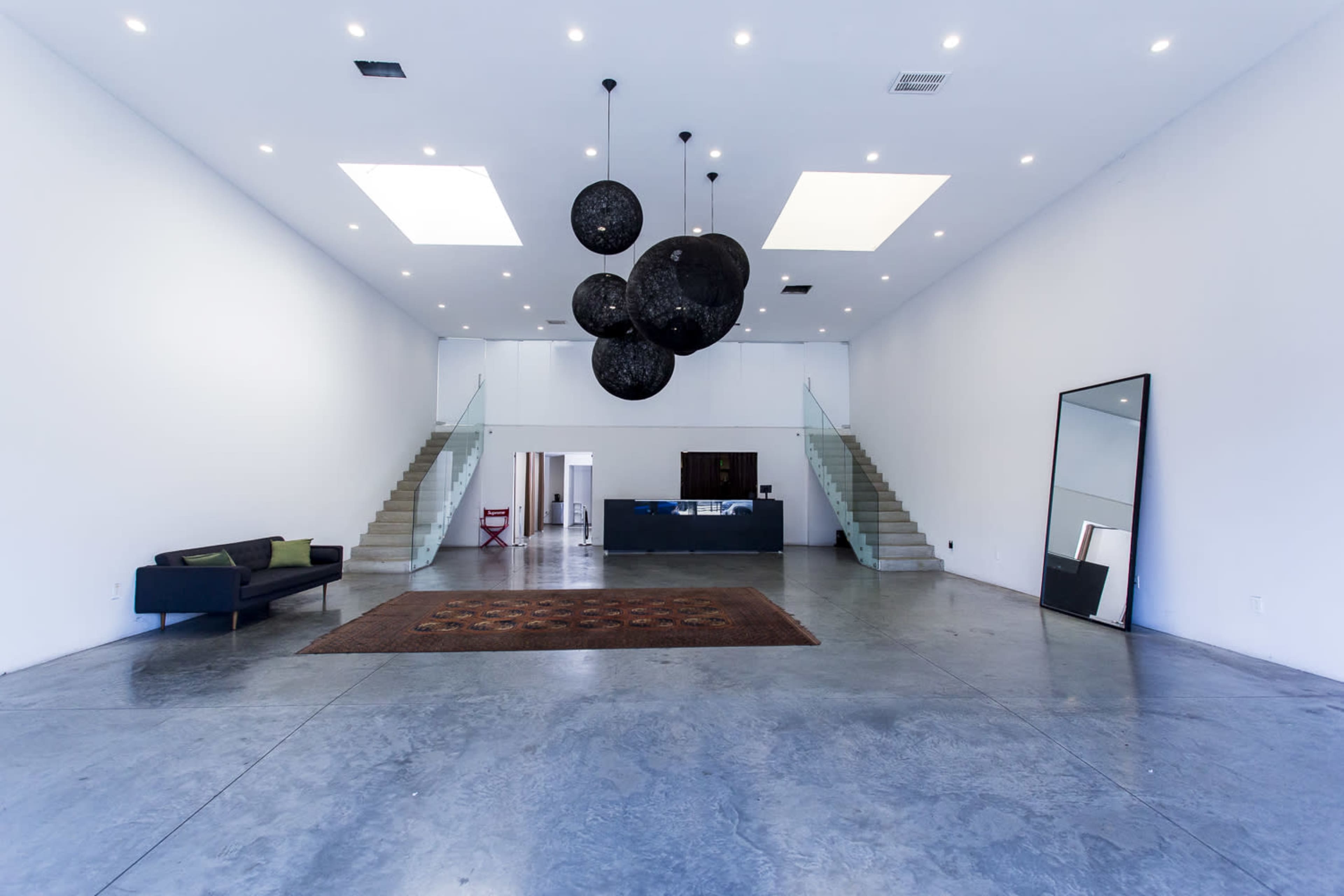 A spacious, minimalist interior features a large open area with concrete flooring, two sets of stairs, a dark rug, a modern sofa, and circular hanging light fixtures.