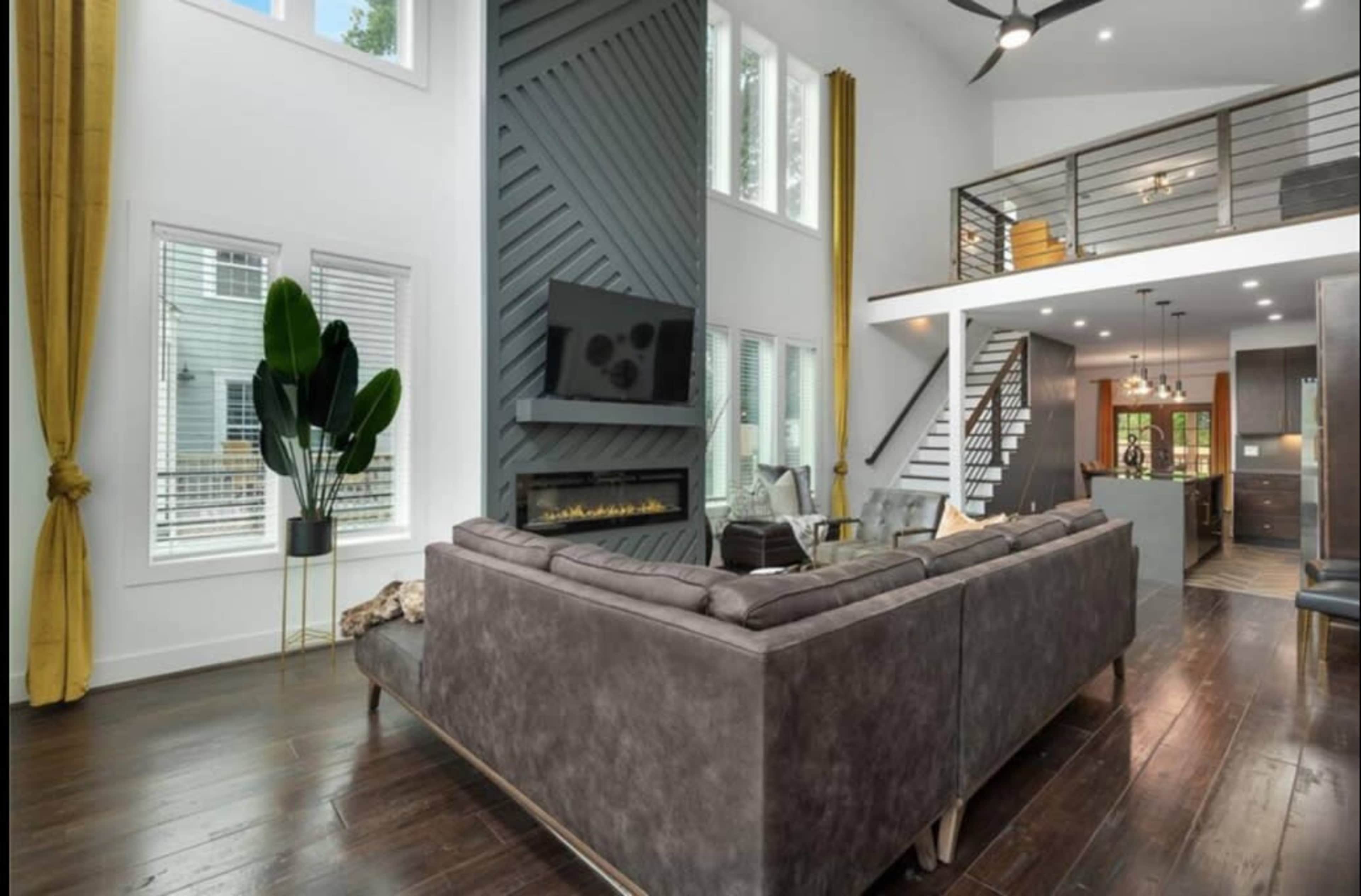 A modern living room features a sectional sofa, a sleek fireplace, large windows with yellow curtains, and a balcony overlooking the kitchen area.