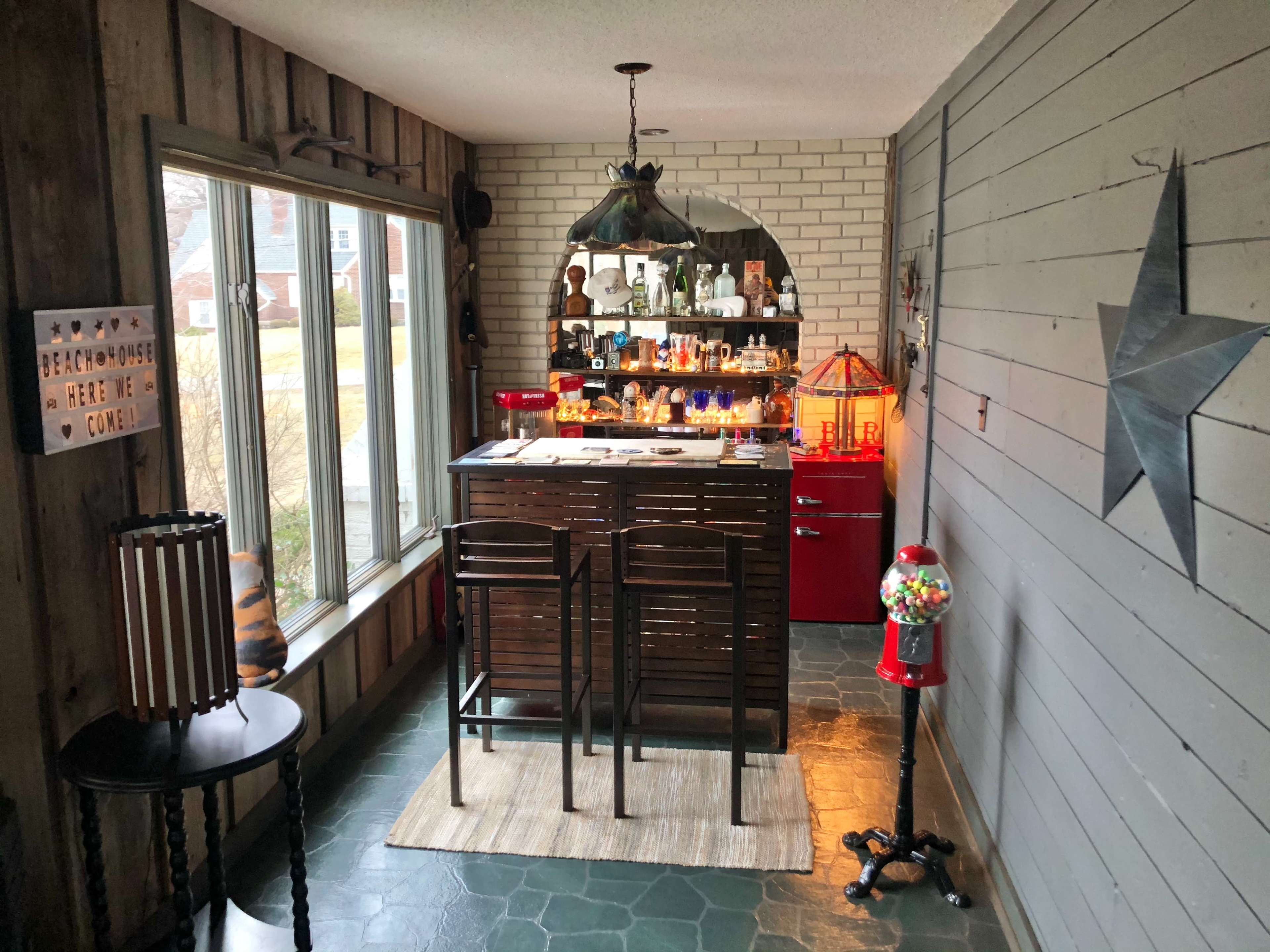 A cozy room features a wooden bar counter with two stools, facing a shelf filled with various collectibles, while a bright red refrigerator stands beside a window.
