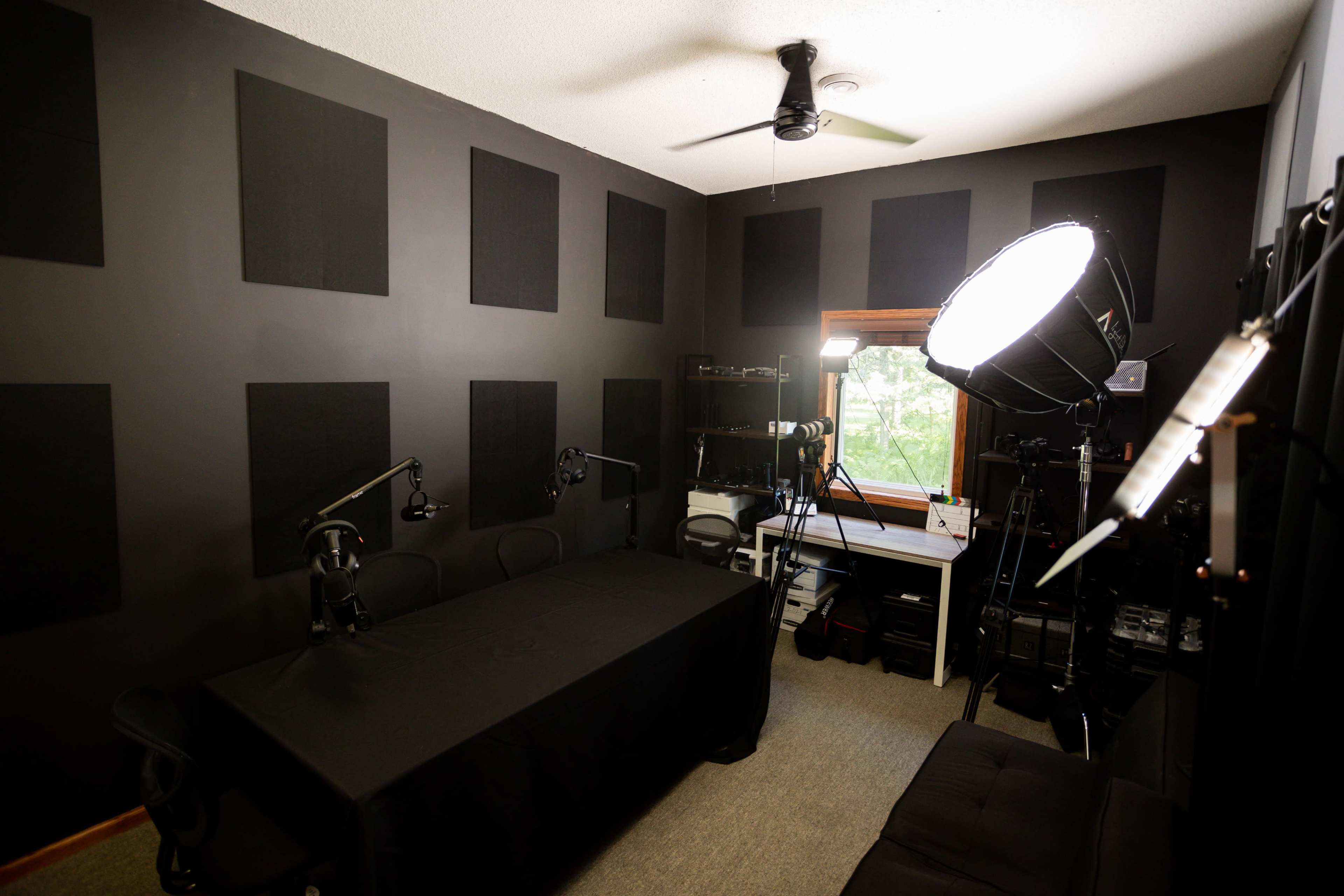 A recording studio with a dark panelled walls, a large table covered with a black cloth, several microphones on stands, and equipment positioned around the room, including a large light source and a window.