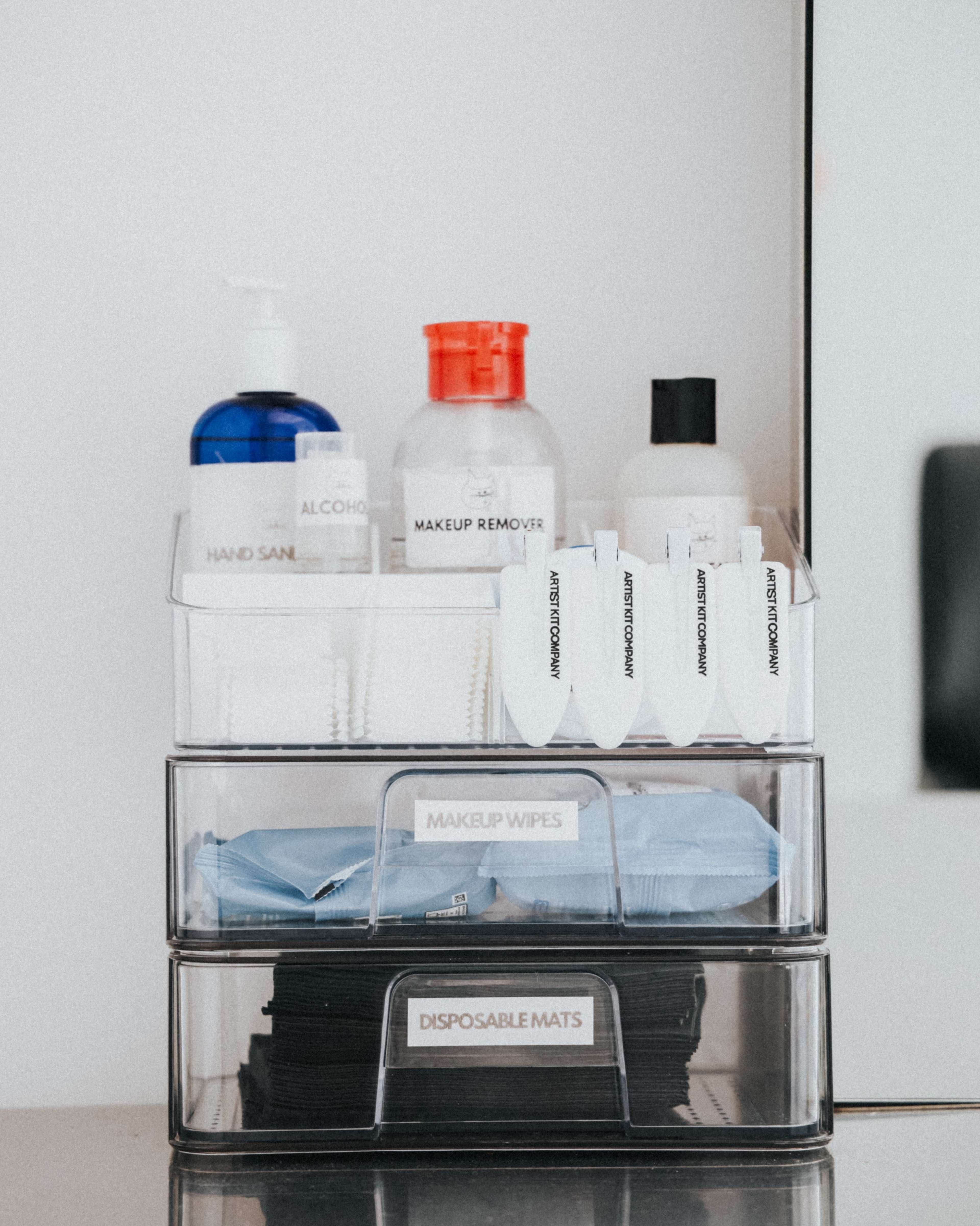 A clear storage container holds various personal care products, including hand sanitizer, makeup remover, and makeup wipes, organized on a counter.