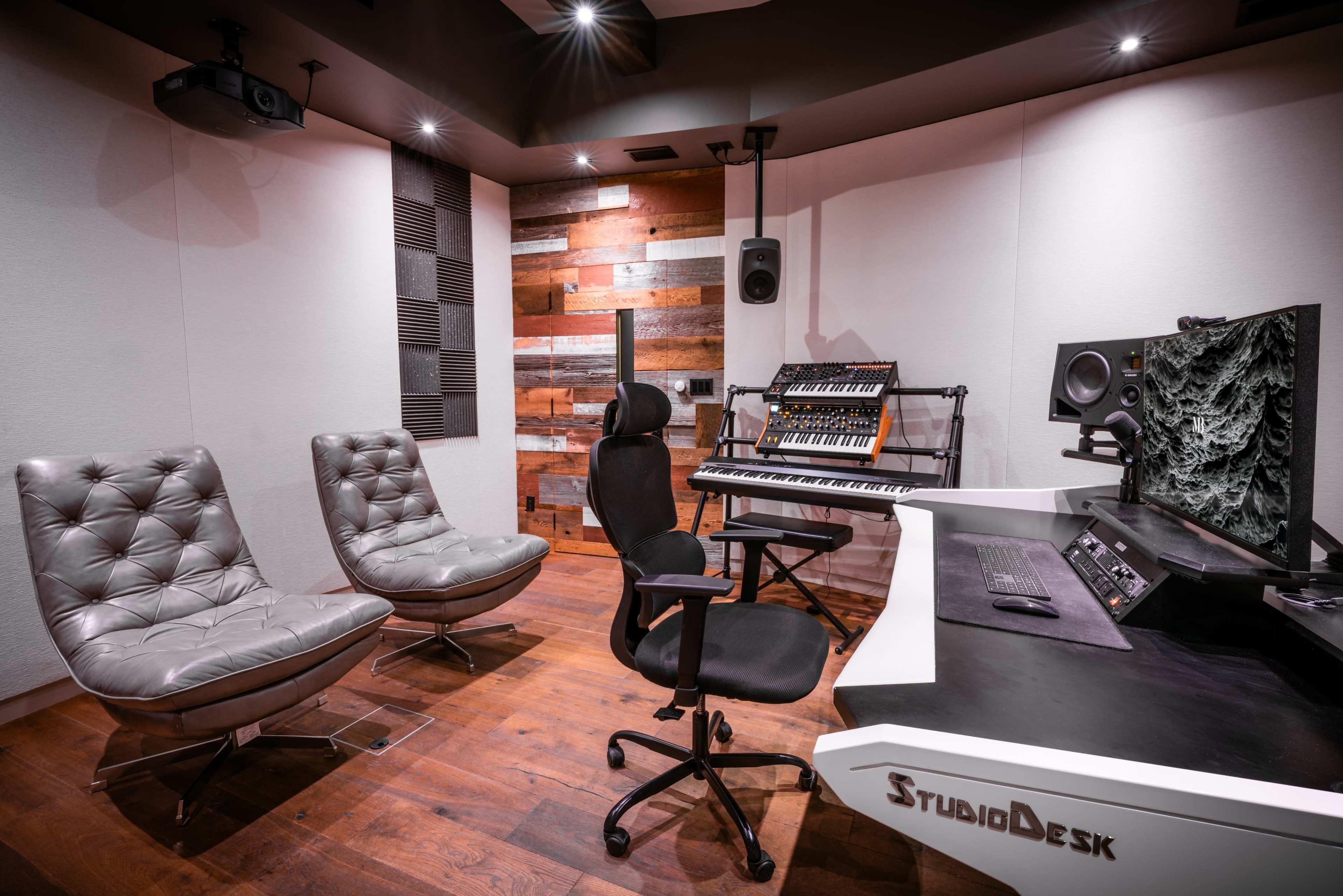 The image shows a modern music studio featuring two stylish lounge chairs, a desk with audio equipment, and a synthesizer against a backdrop of wooden paneling.