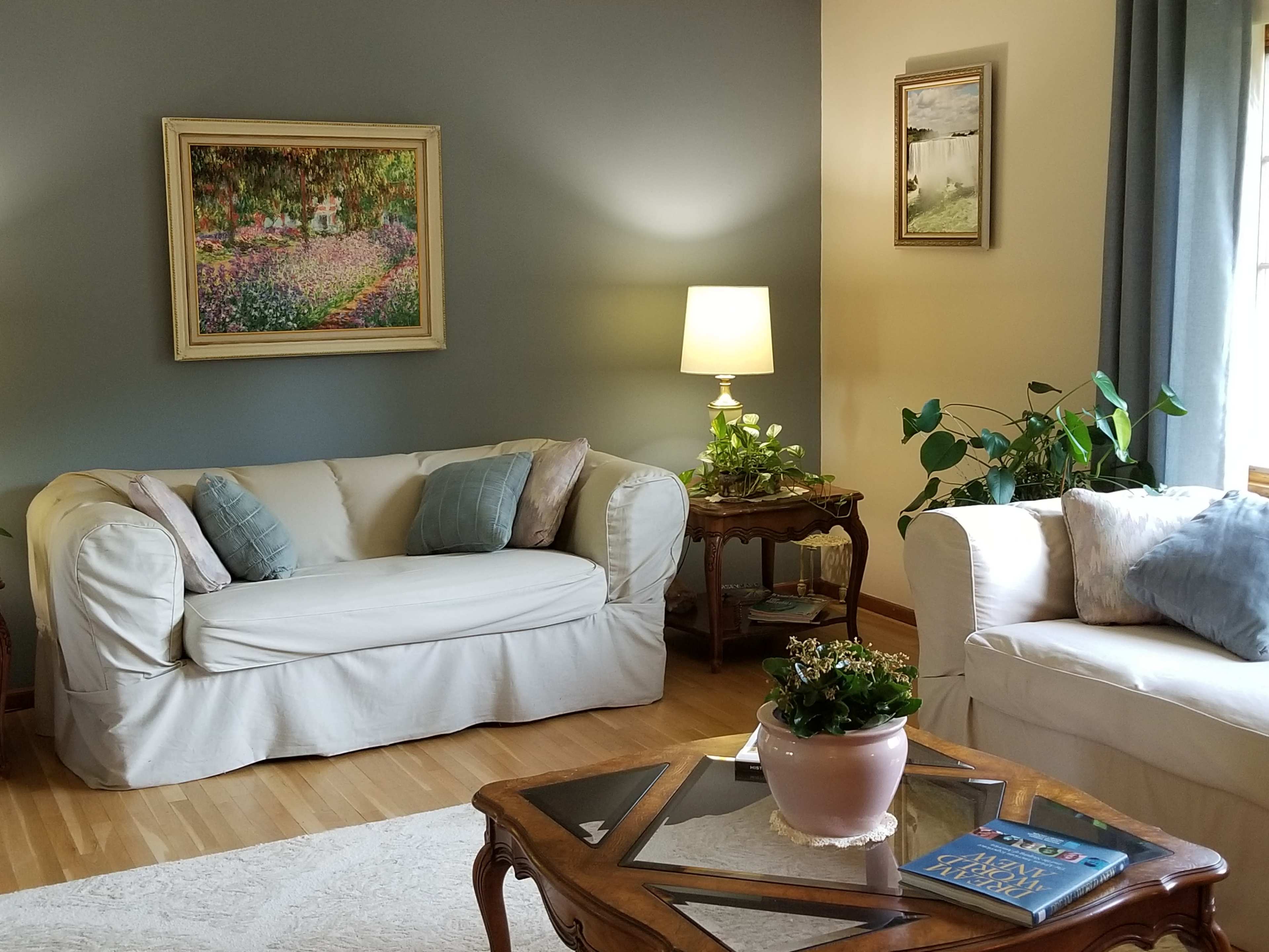 A cozy living room features a light-colored sofa, a wooden coffee table with a book, and a lamp next to a potted plant by the window.