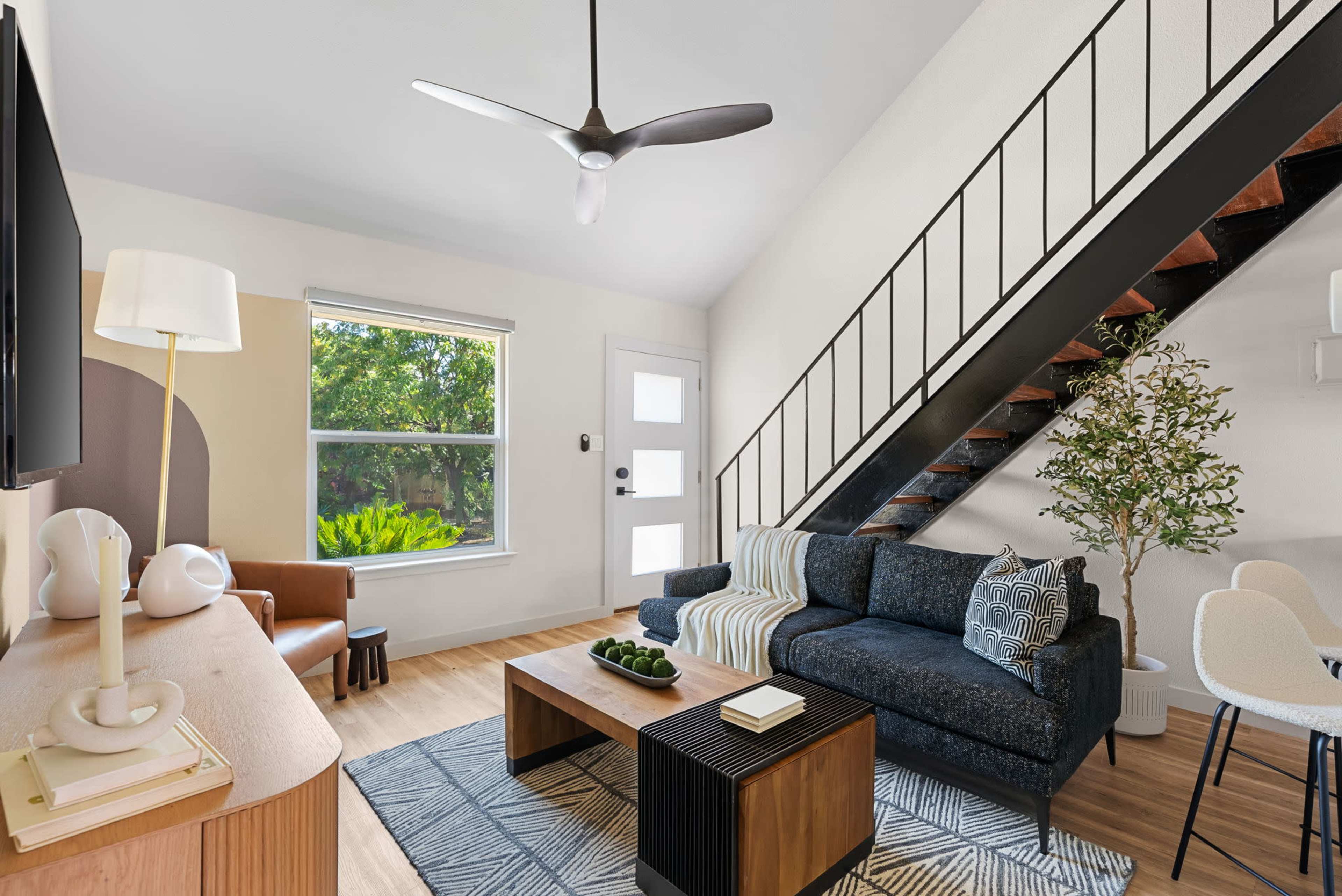 The image shows a modern living room with a sectional couch, a wooden coffee table, a staircase, and a large window letting in natural light.