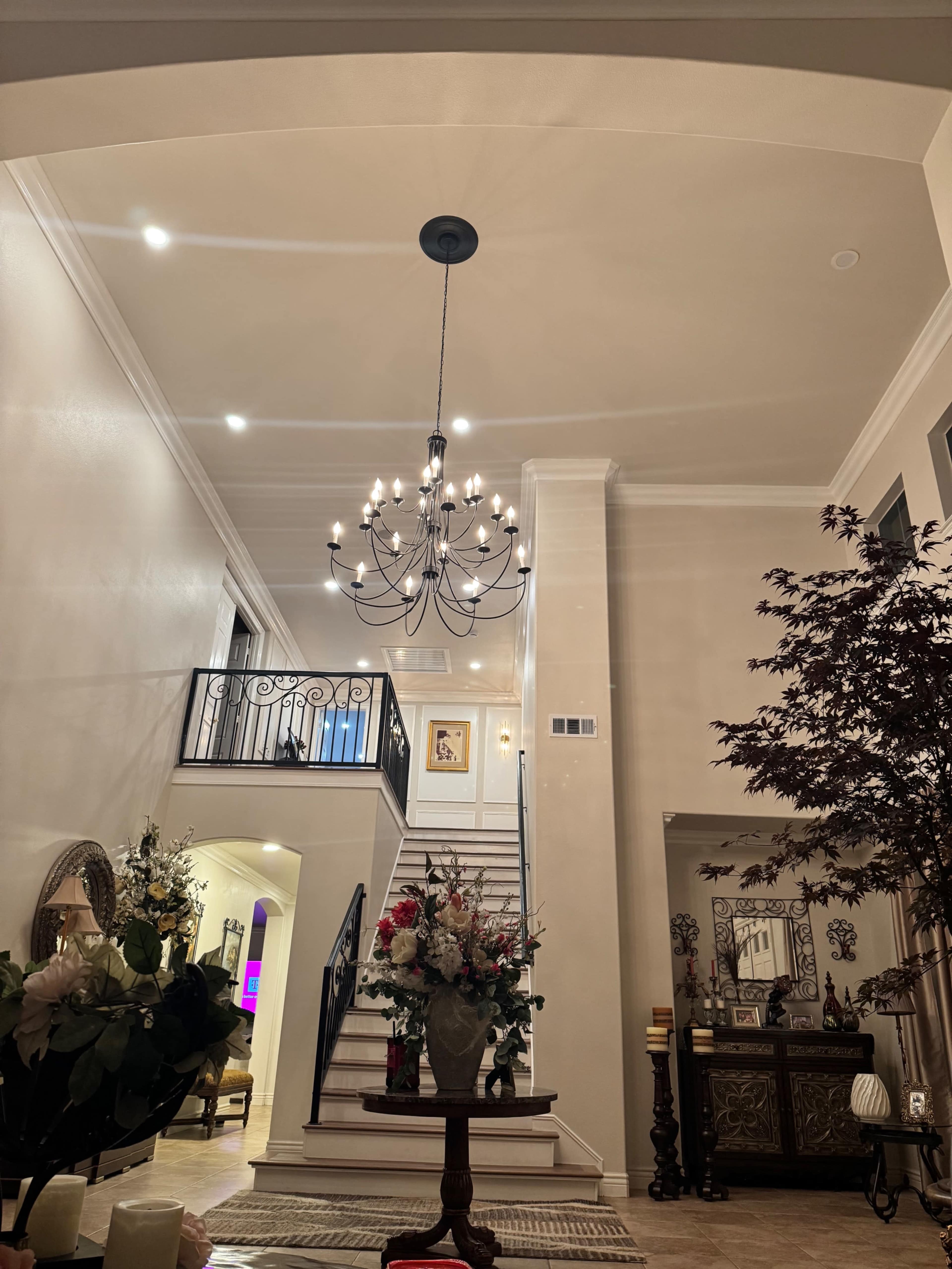 The image shows a spacious entryway with a chandelier, a staircase leading to an upper floor, and decorative plants and flowers in vases.
