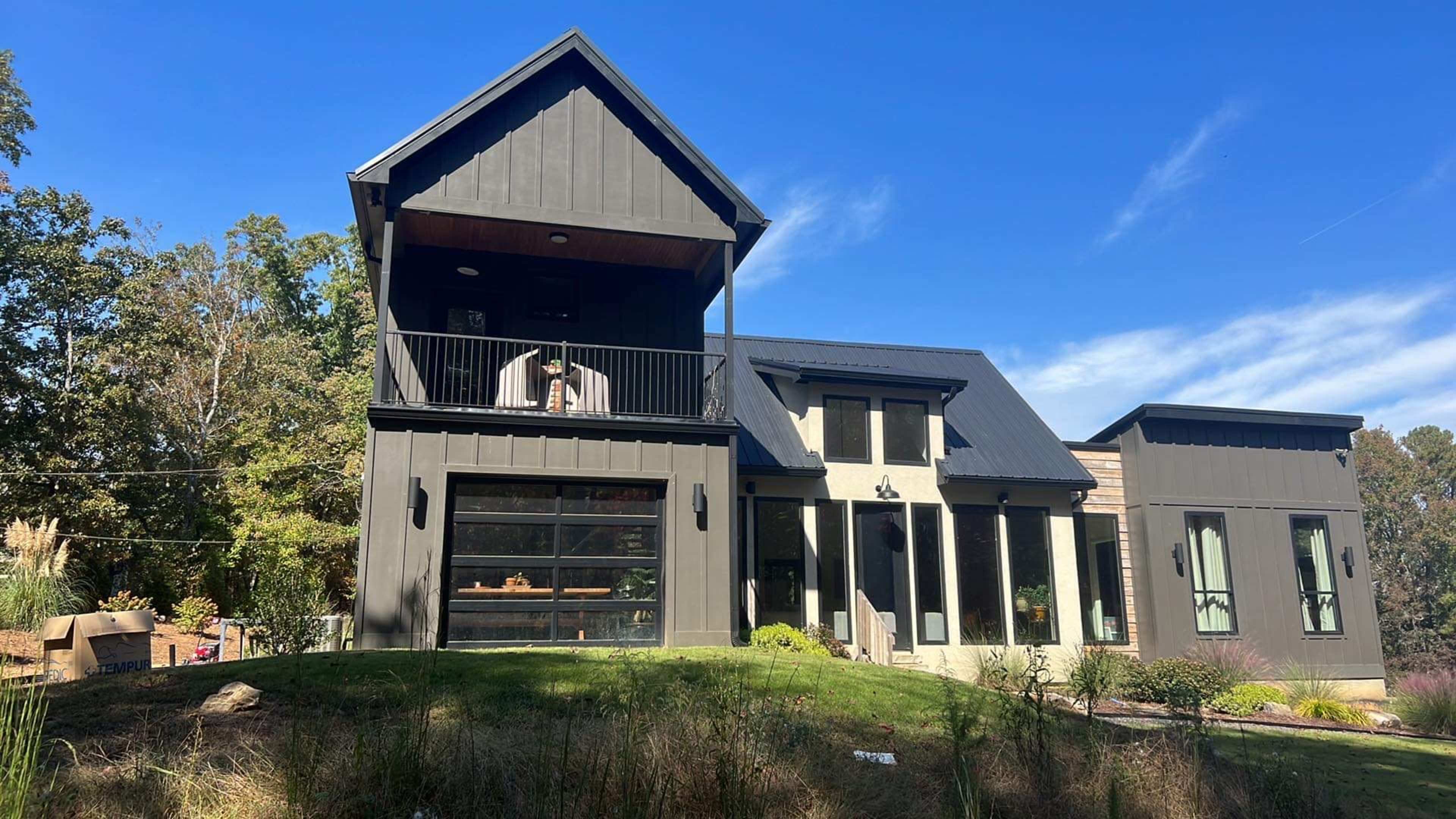 A modern black-clad house features a prominent balcony and large windows, surrounded by greenery and trees.