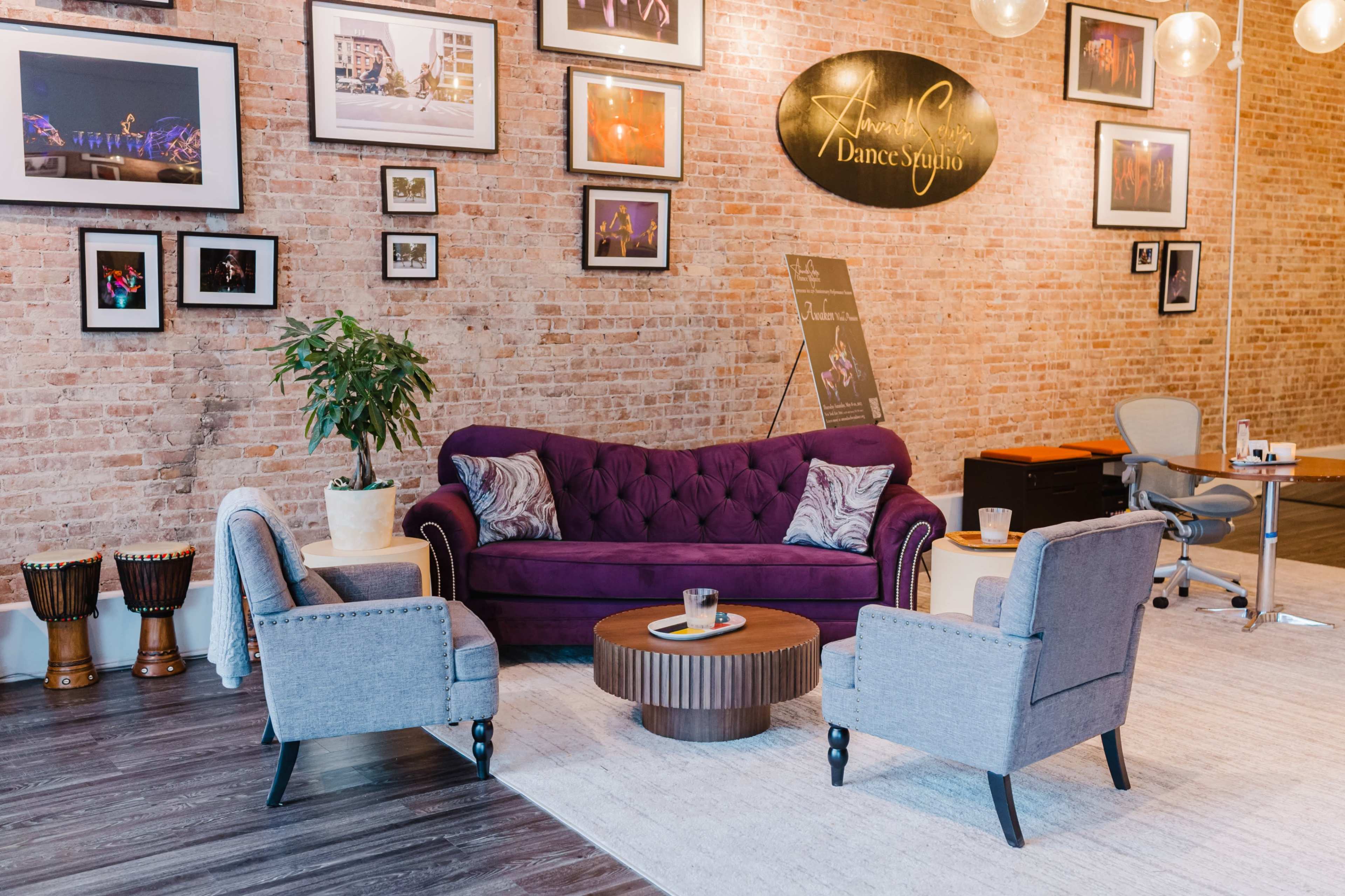 The image shows a dance studio waiting area with a purple tufted sofa, two gray armchairs, a round coffee table, and a plant, against a backdrop of exposed brick walls and framed photos.
