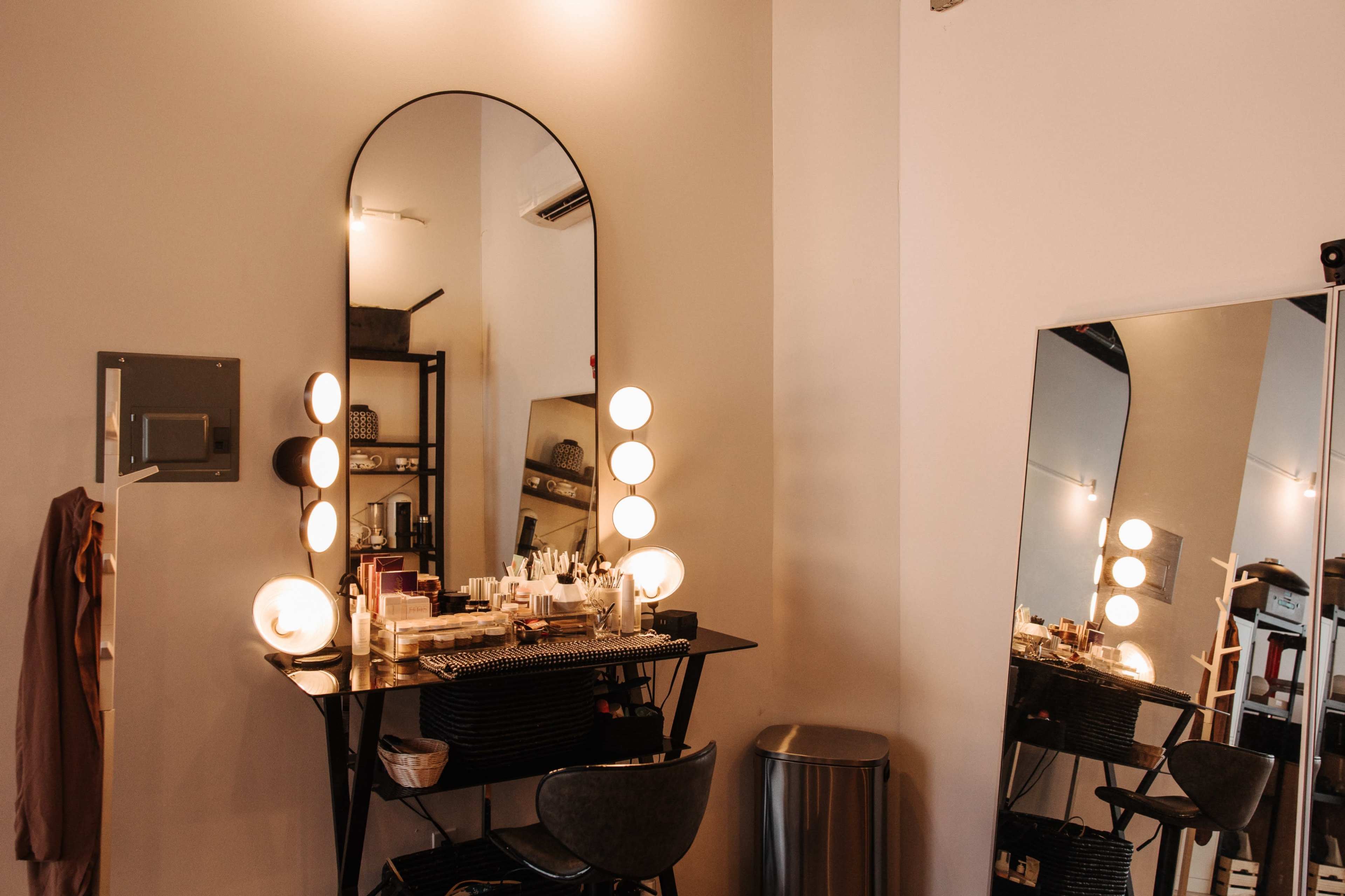The image shows a modern vanity area with a large, oval mirror surrounded by light bulbs, a black table with various beauty products, and a tall mirror reflecting the space.