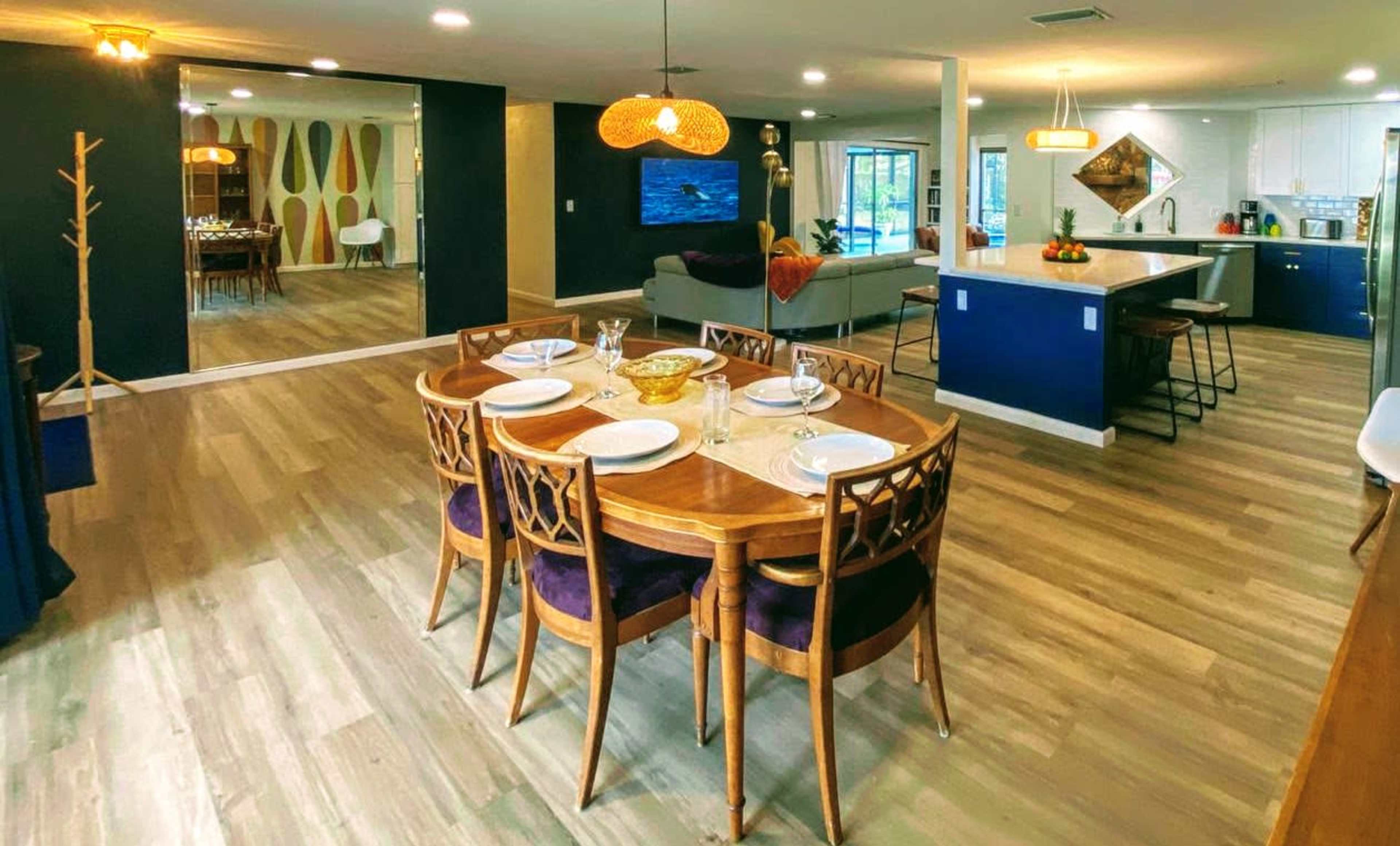 A modern, open-concept dining and living area with a wooden dining table set for four, a kitchen with a central island, and a large wall-mounted television.