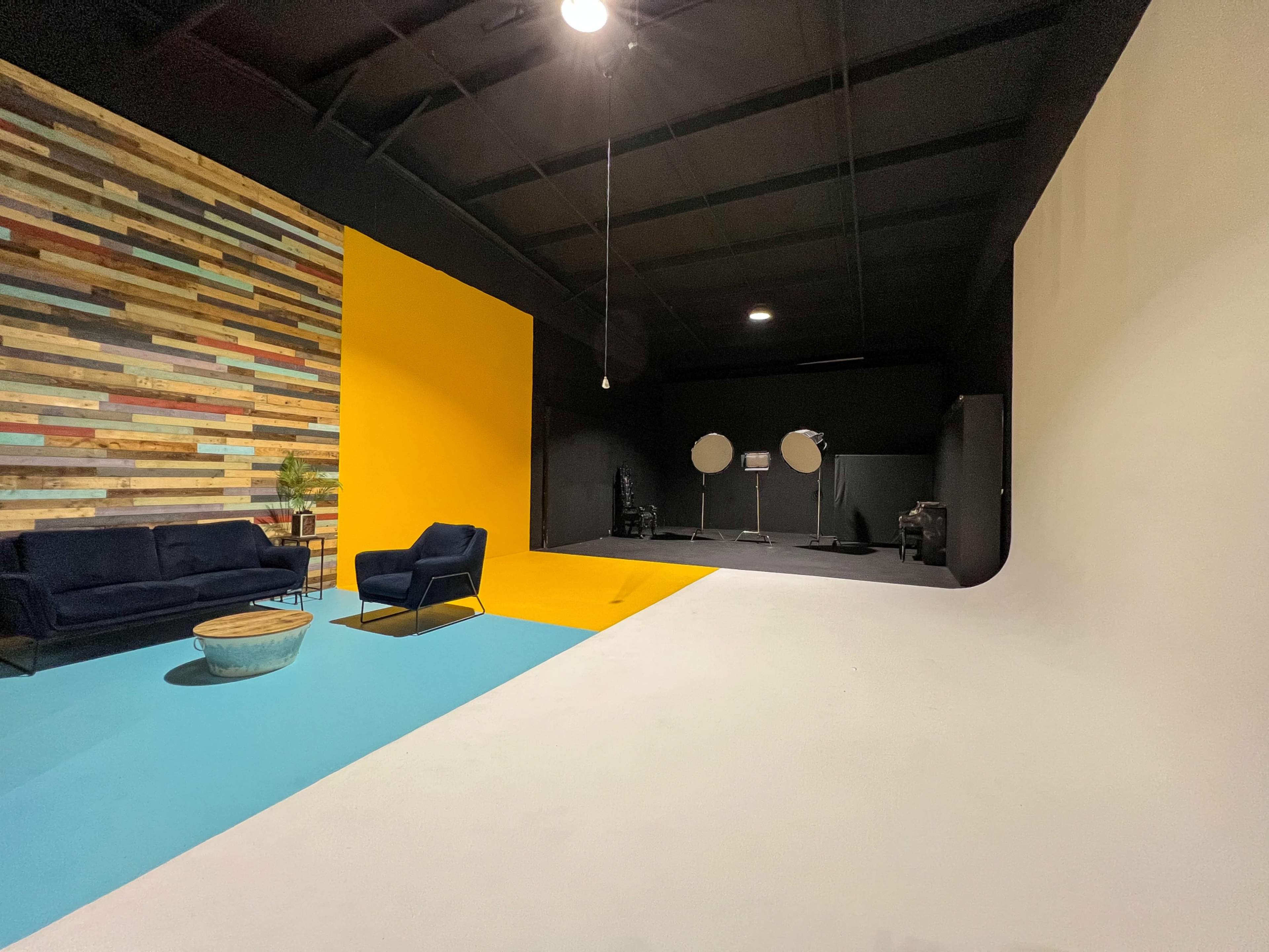 The image shows a spacious studio with a textured wooden accent wall, a bright yellow section, a light blue area, and a white backdrop, featuring a sofa and standing equipment.