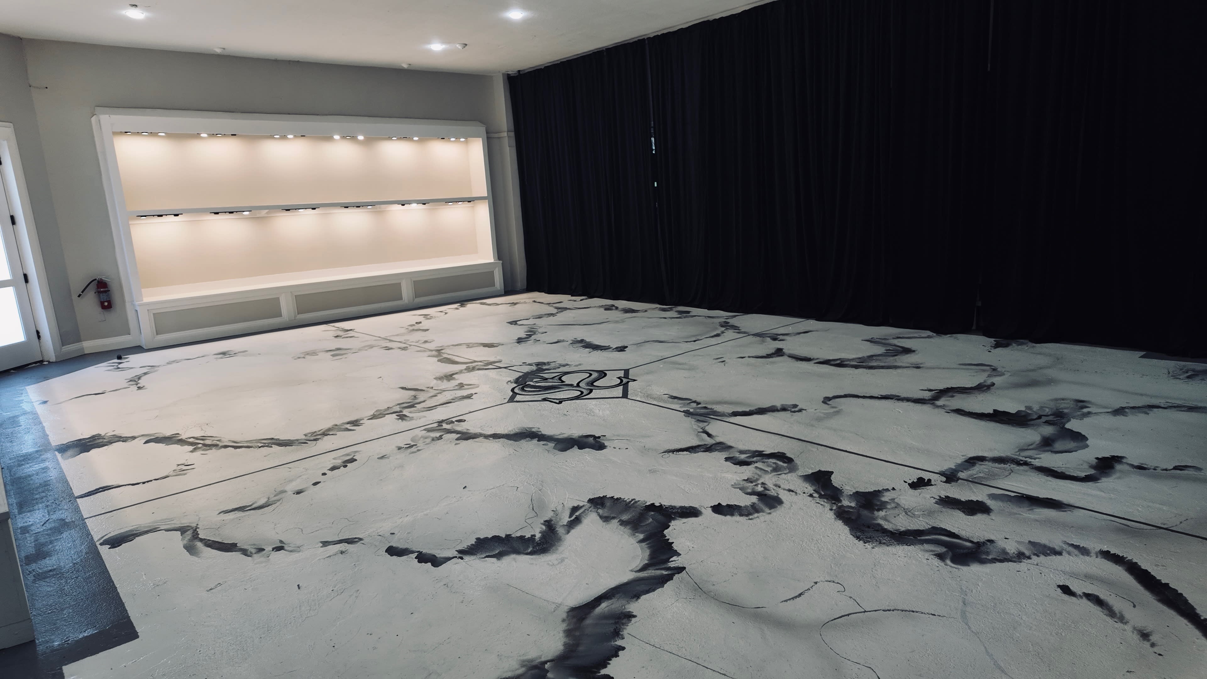 The scene features a large, empty room with a textured, cracked floor design and black curtains along one wall.