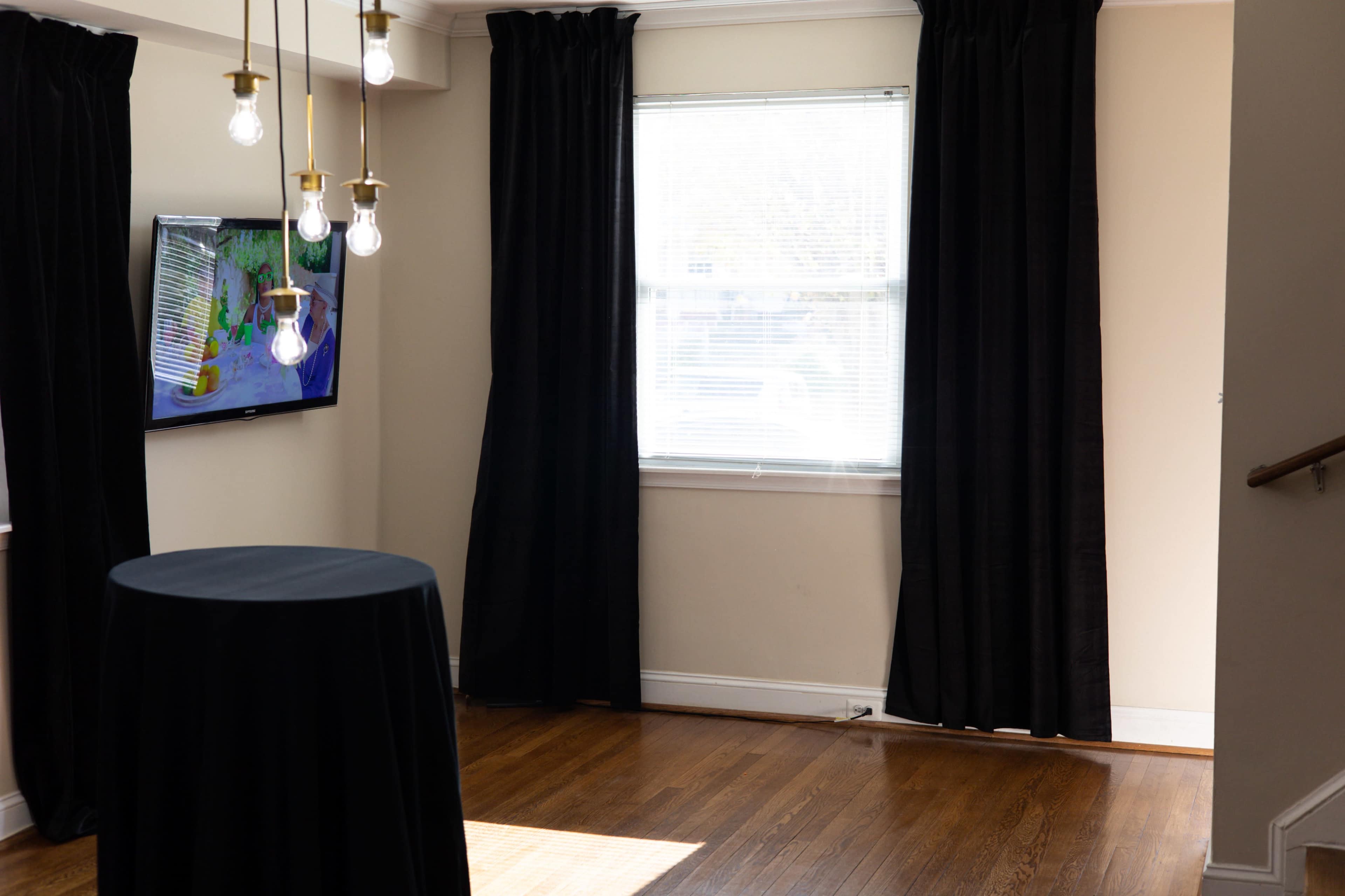 The image shows a room with hardwood flooring, a black table in the center, and curtain-covered windows, alongside a television on the wall displaying colorful graphics.