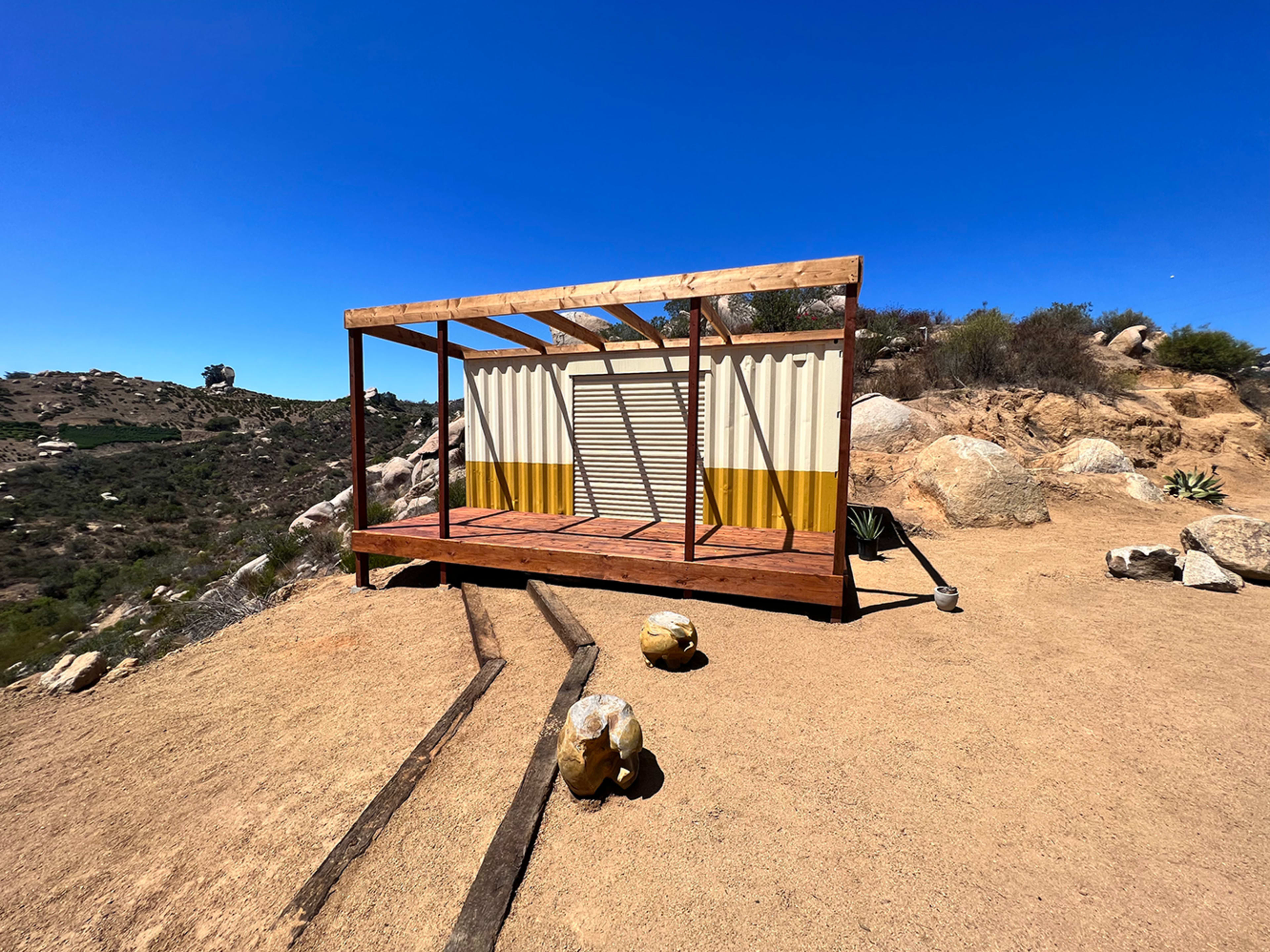 A wooden structure with a corrugated metal container on a rocky, sandy landscape under a clear blue sky.