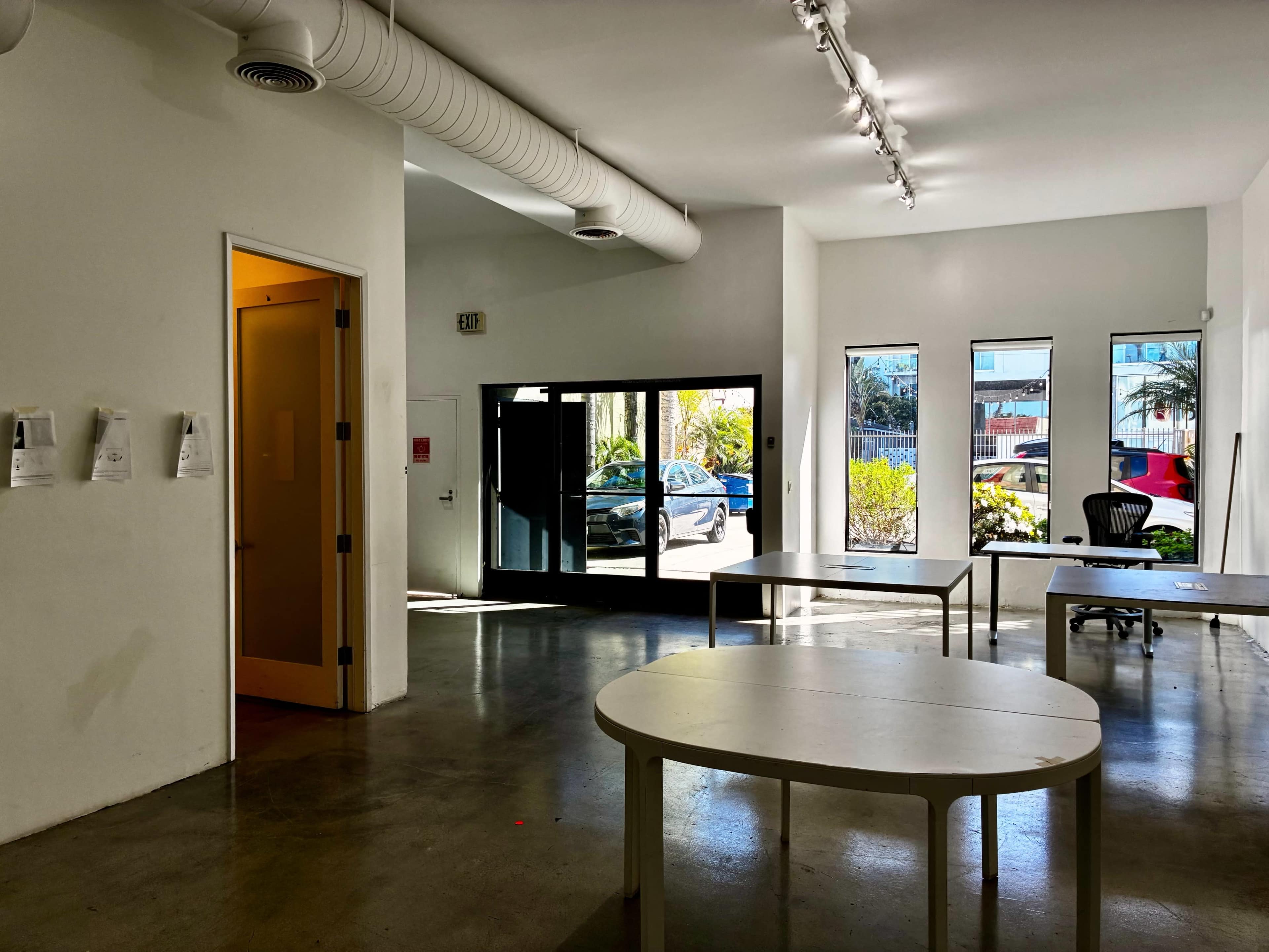 The image shows an interior space with white walls, large windows allowing natural light, and several white tables arranged in the room.