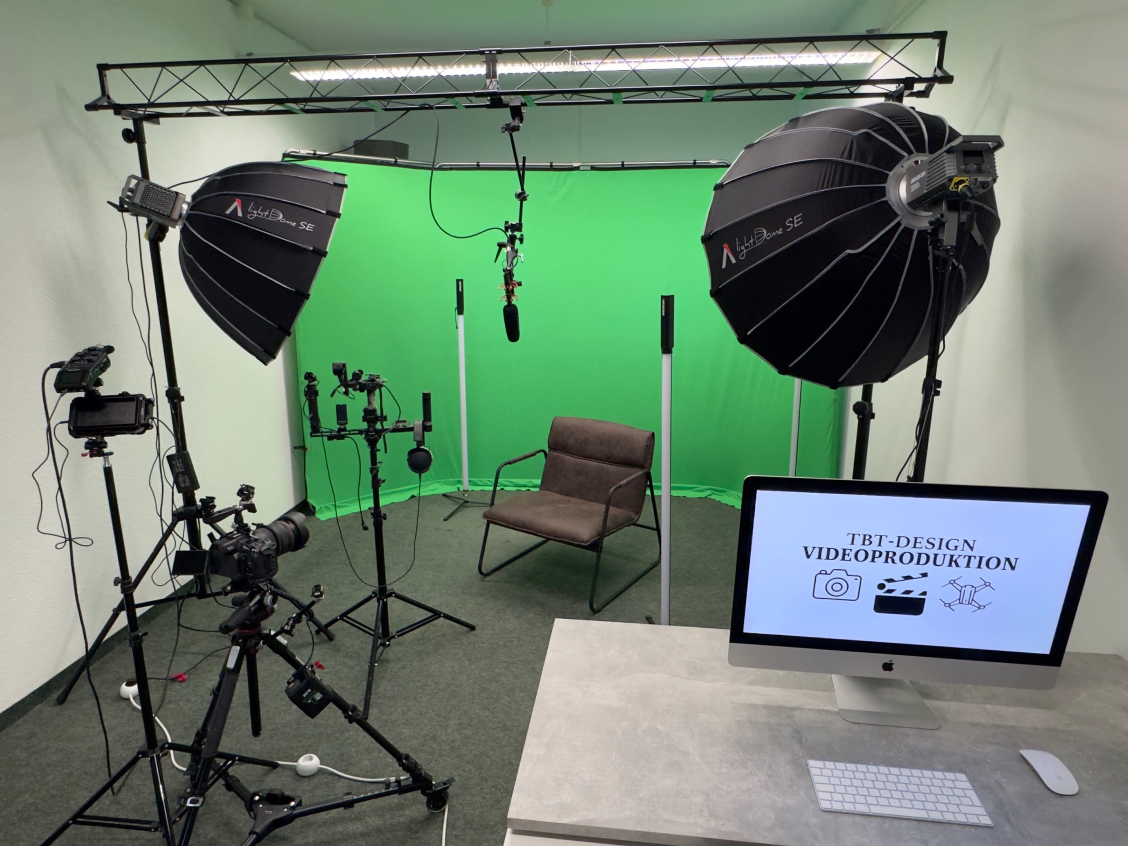 The image shows a video production setup with a green screen, lighting equipment, a camera on a tripod, a microphone, an empty chair, and a computer displaying a design screen.