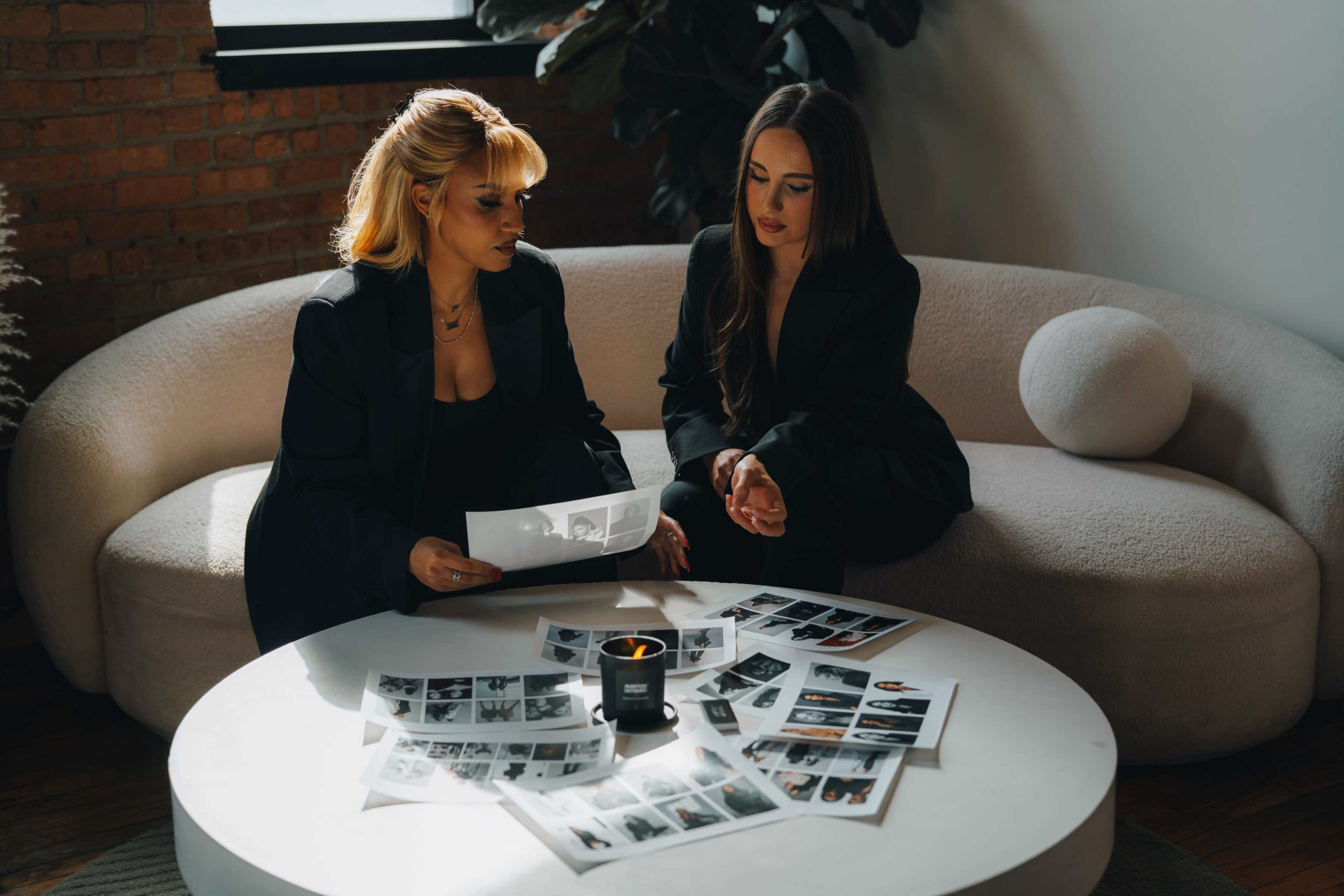 Two women in black blazers are seated on a light-colored couch, examining photo prints arranged on a white table with a candle in the center.