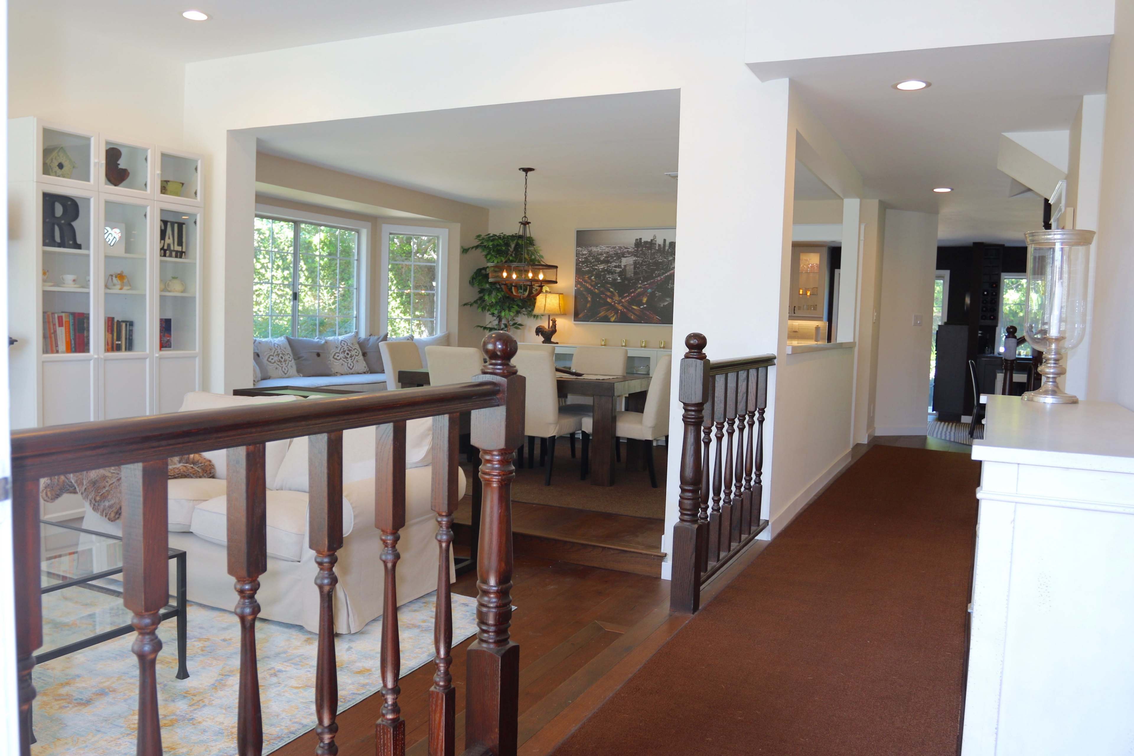 The image shows a well-lit interior space featuring an open floor plan that connects a living area with a dining space, separated by a staircase and a hallway.