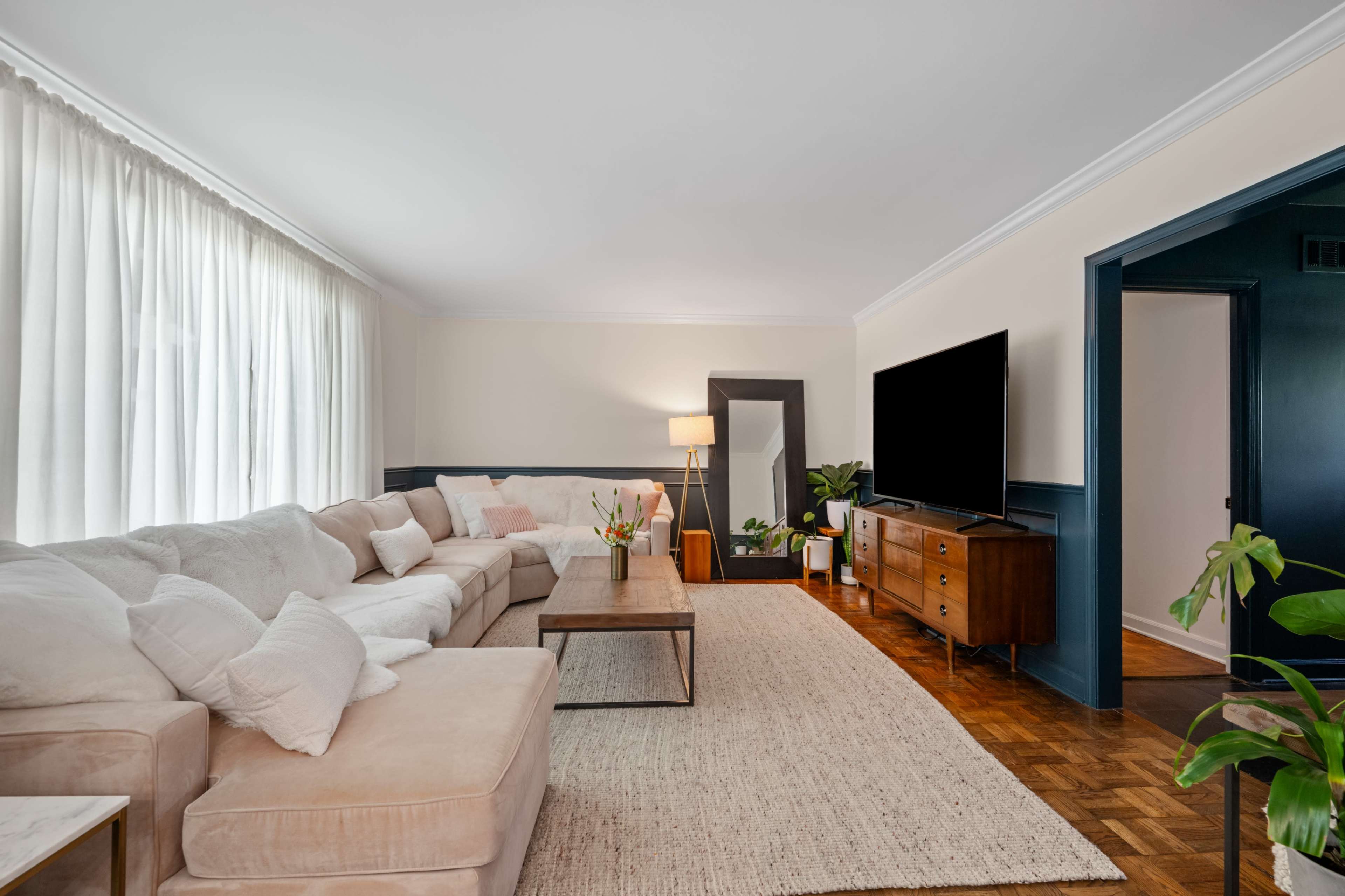 A bright living room features a large beige sectional sofa, a wooden coffee table, and a TV stand with a flat-screen television, complemented by natural light filtering through white curtains.