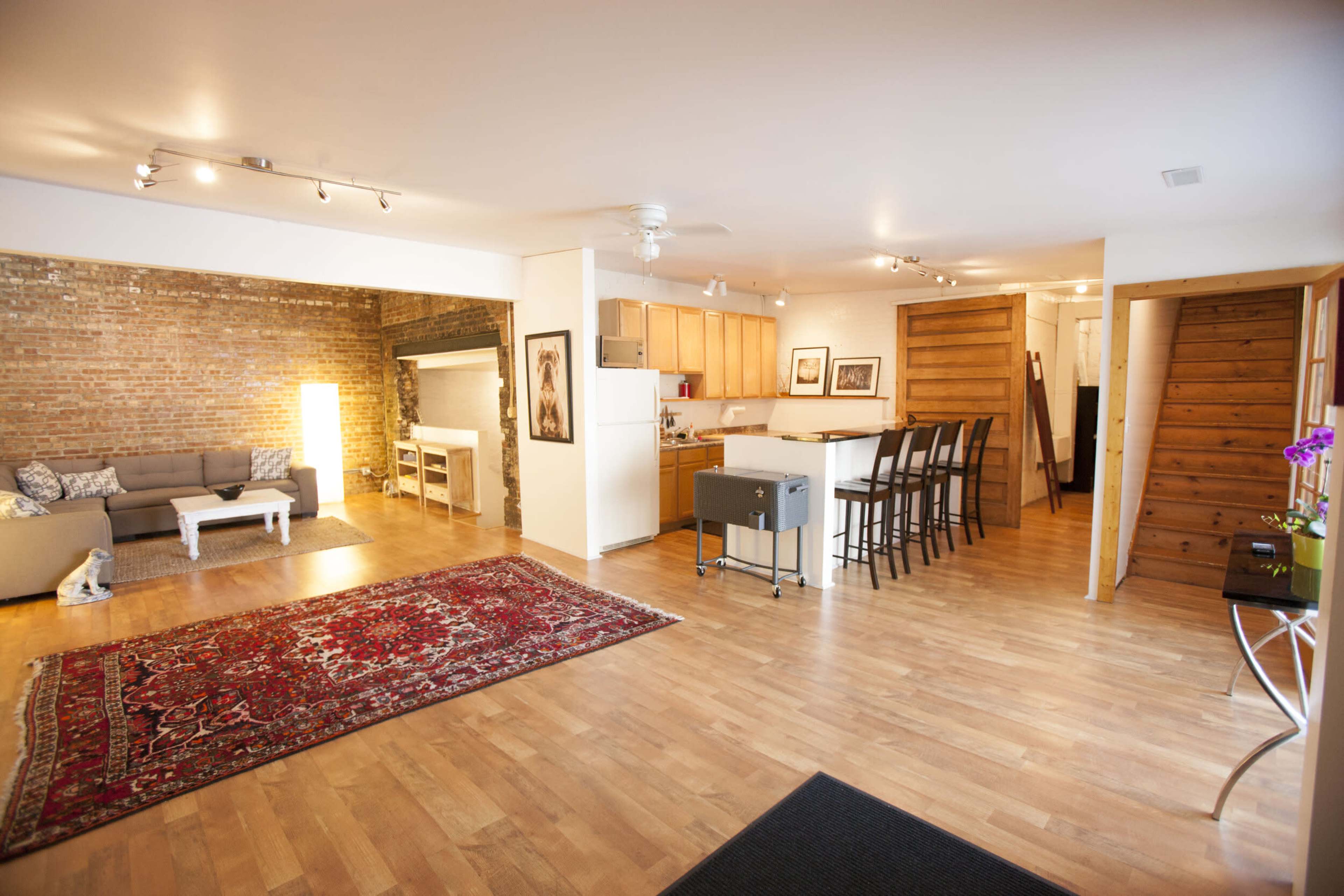 A spacious living area with hardwood floors, a large area rug, a sectional sofa, and a kitchen with wooden cabinets in an open-concept design.