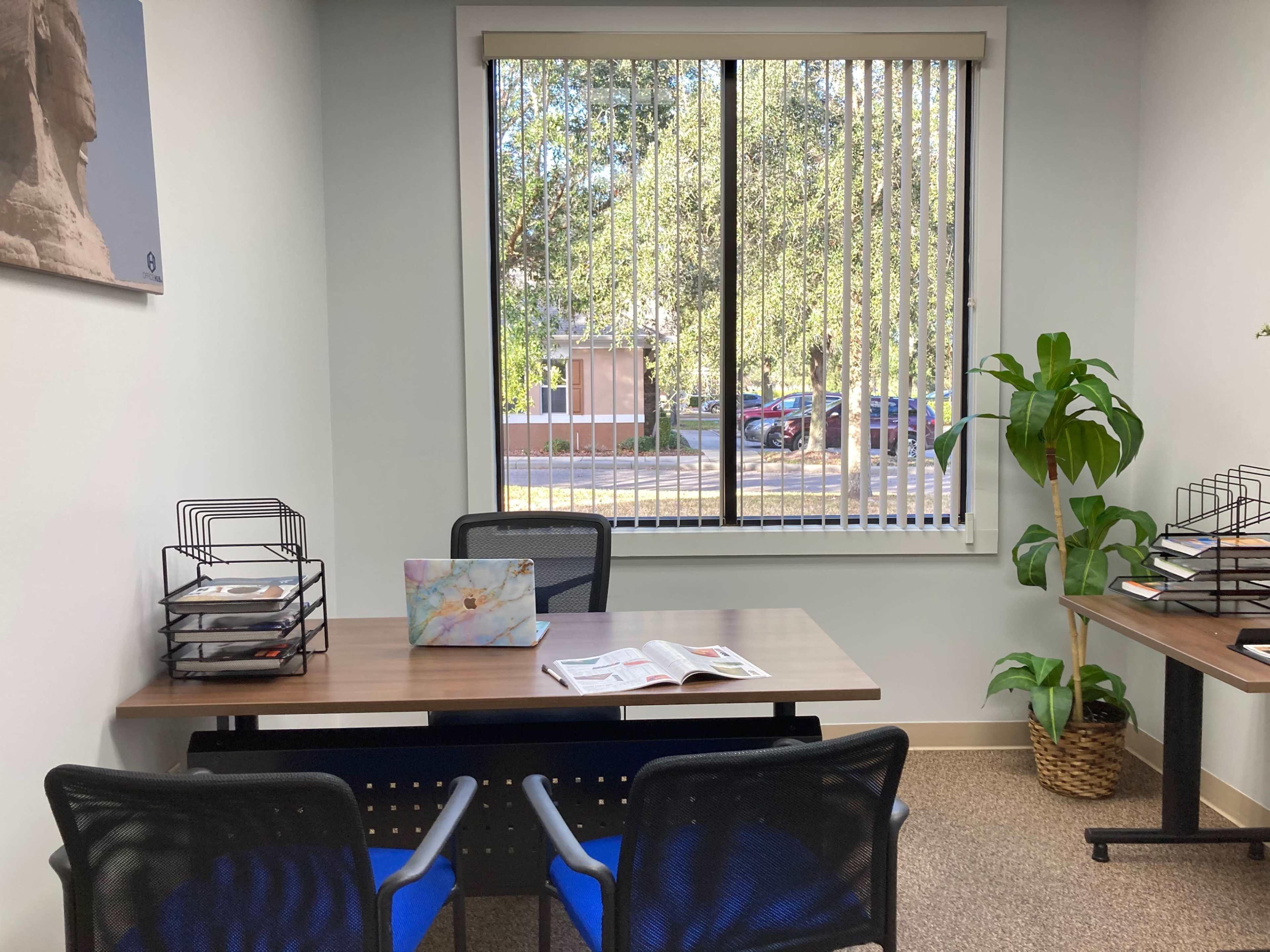 A small office with two empty desks, a plant, and a large window that overlooks a parking lot and trees.